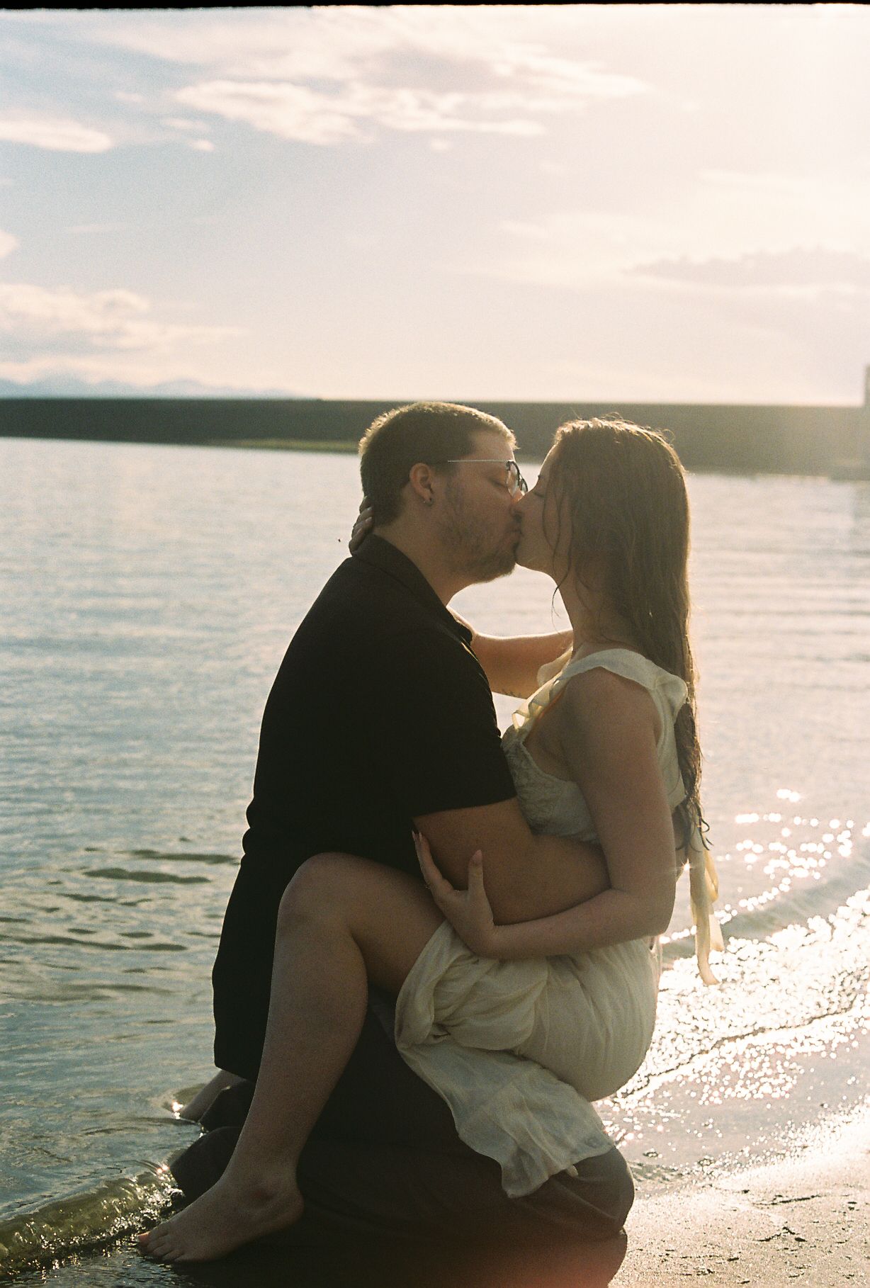 Couple sharing a kiss while sitting together on a lakeside shore during a beautiful golden sunset engagement session.