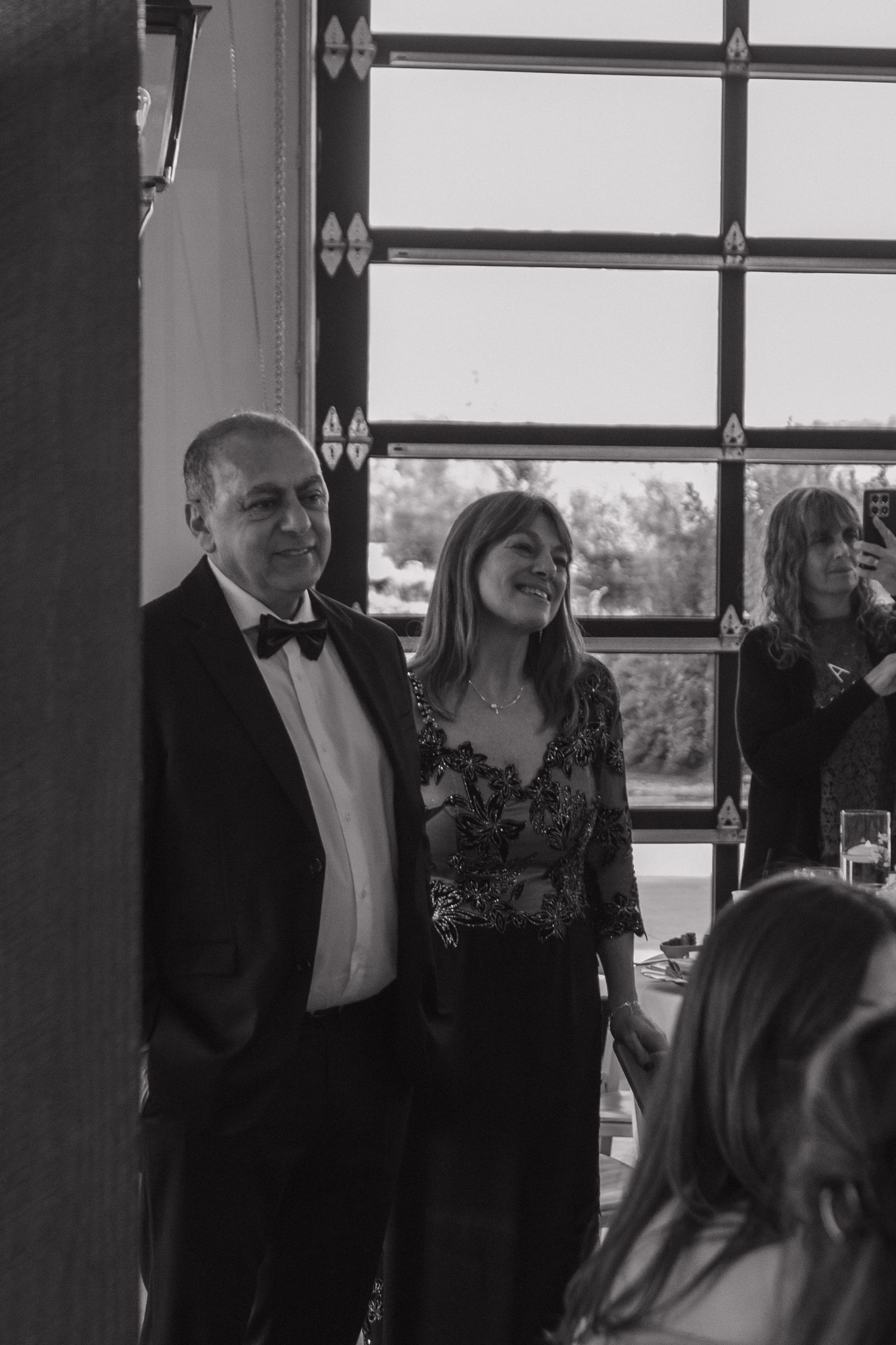 Wedding reception scene with guests gathered by large windows in formal attire.