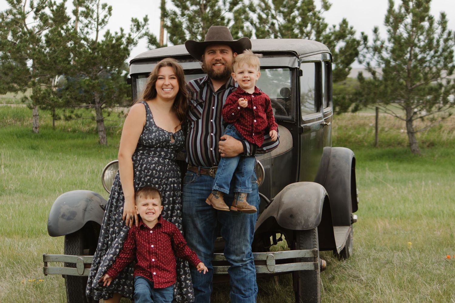 Family portrait in western attire standing by an antique car in a grassy field with pine trees.