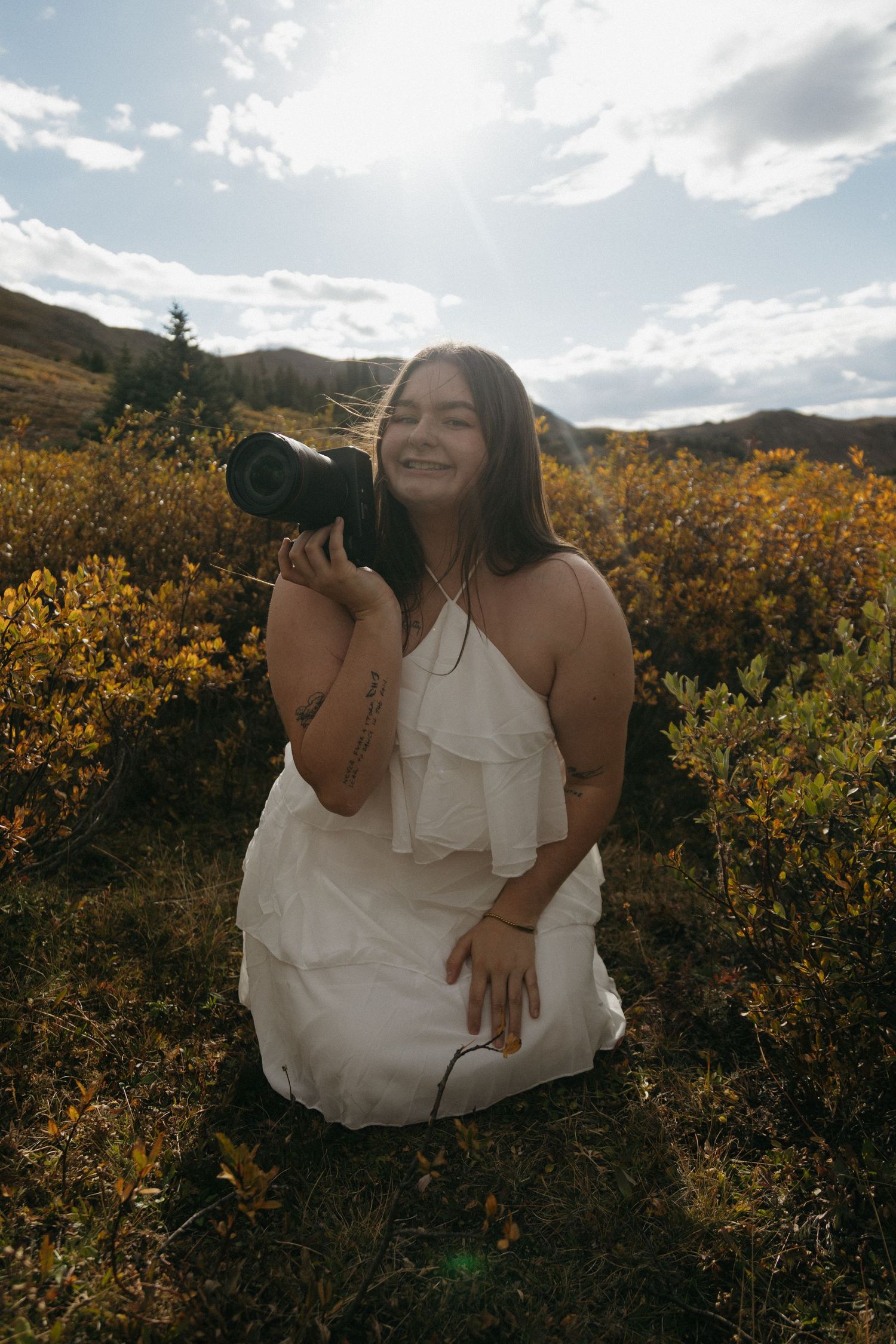 Woman in white dress kneeling in autumn field holding camera, smiling in golden sunlight with mountains behind.