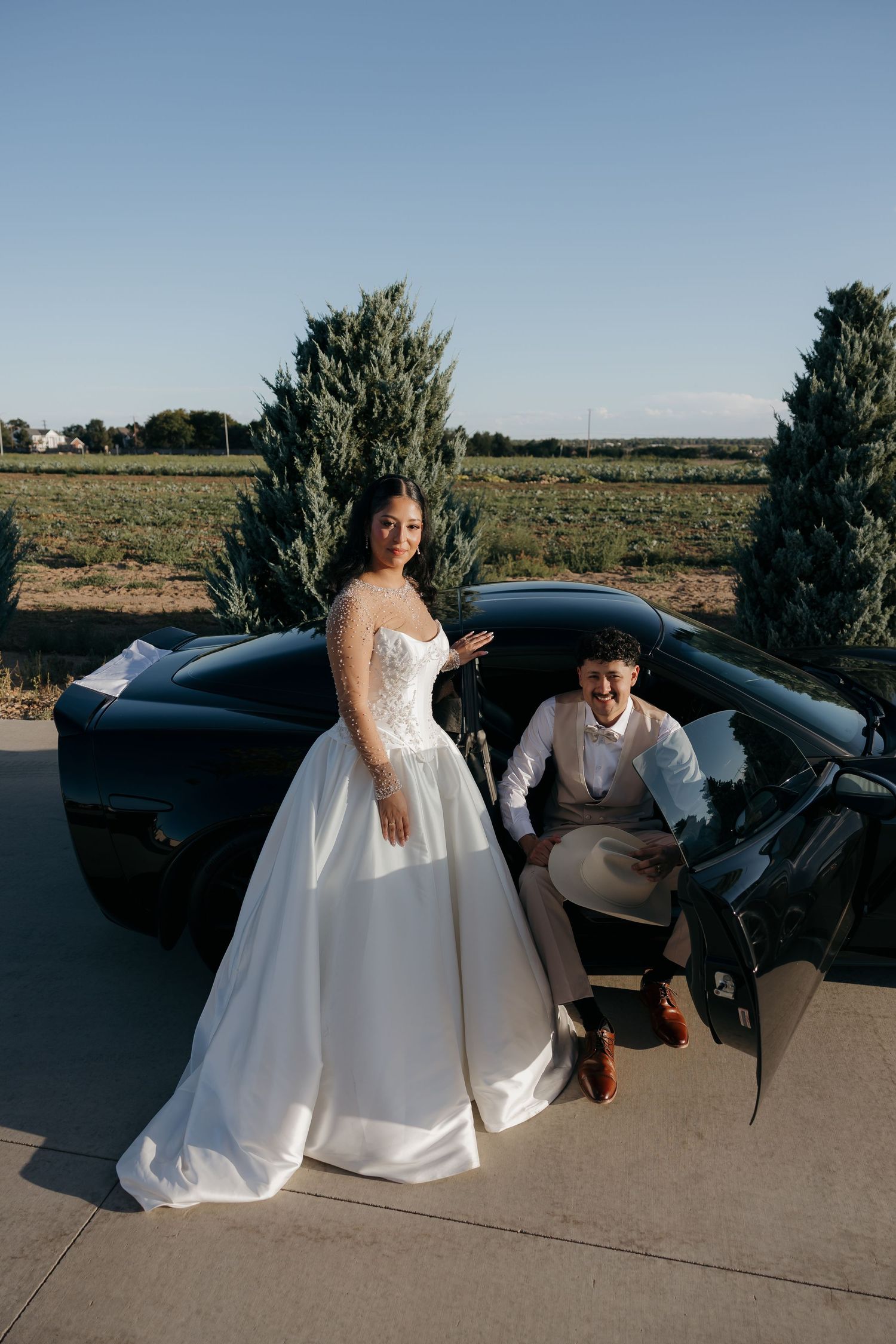 Elegant bride in white ballgown stands beside groom near a black sports car at sunset.