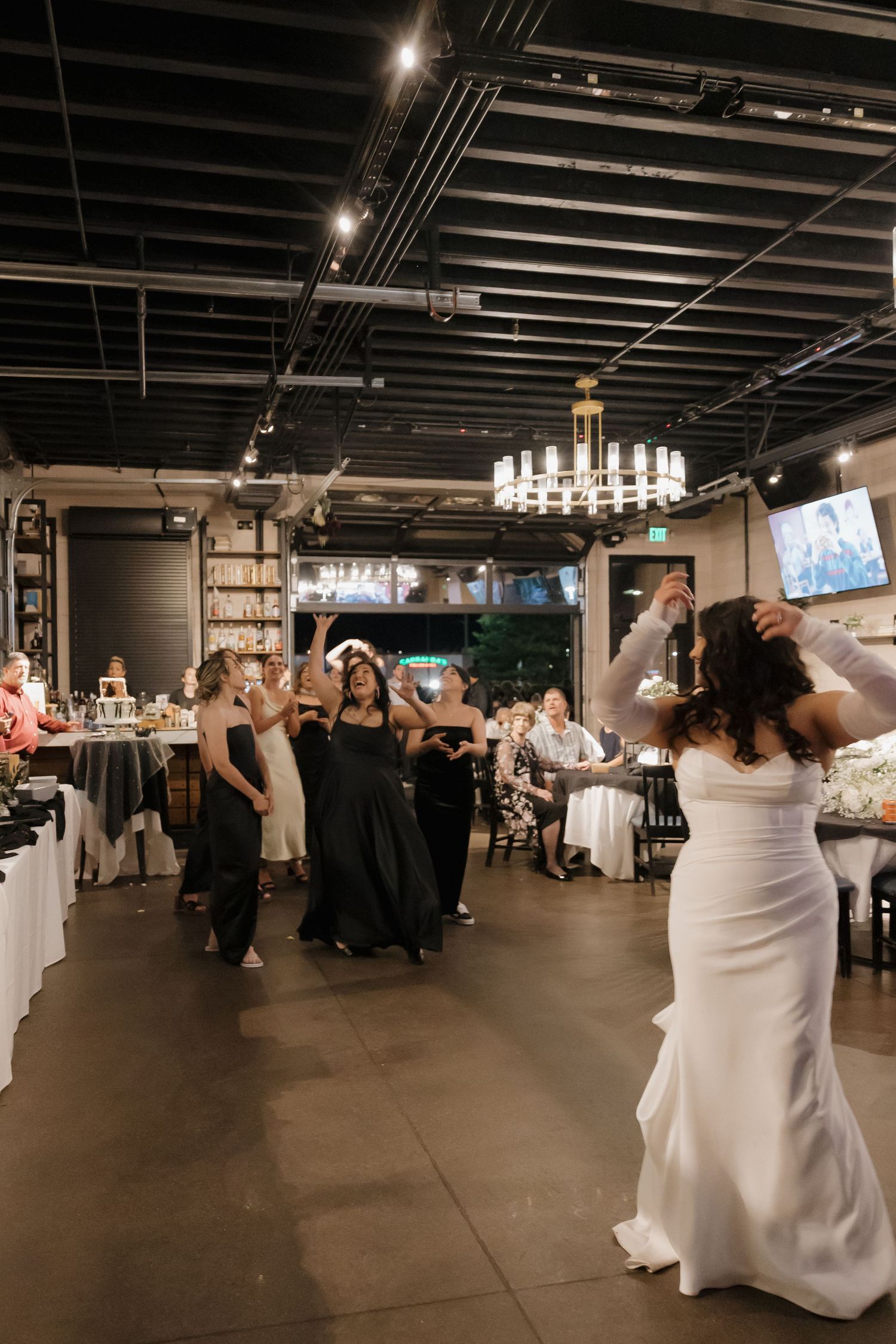 Bride in white gown tosses bouquet to excited guests in elegant industrial wedding reception venue.
