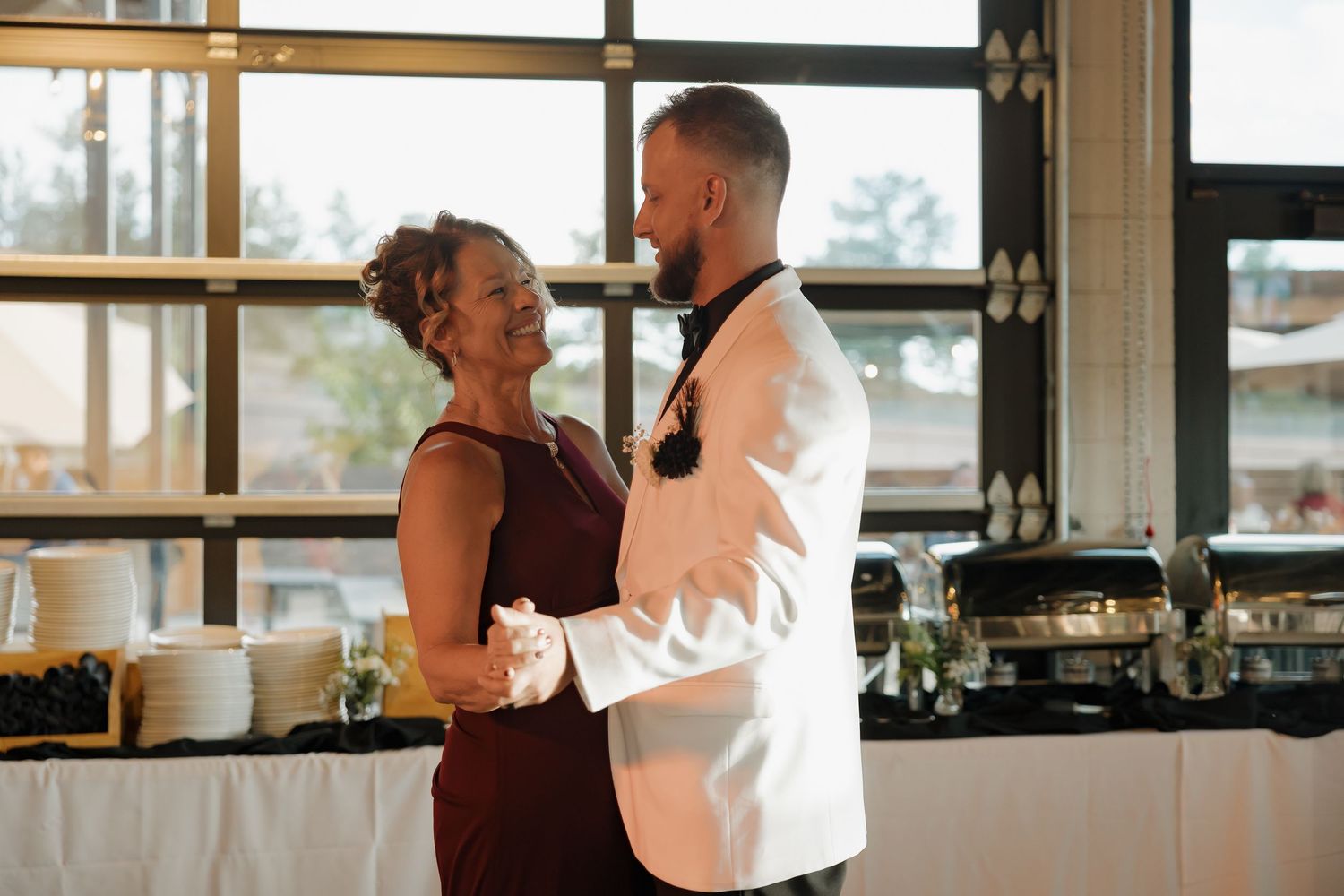 Groom in white jacket shares a mother-son dance at an indoor wedding reception.