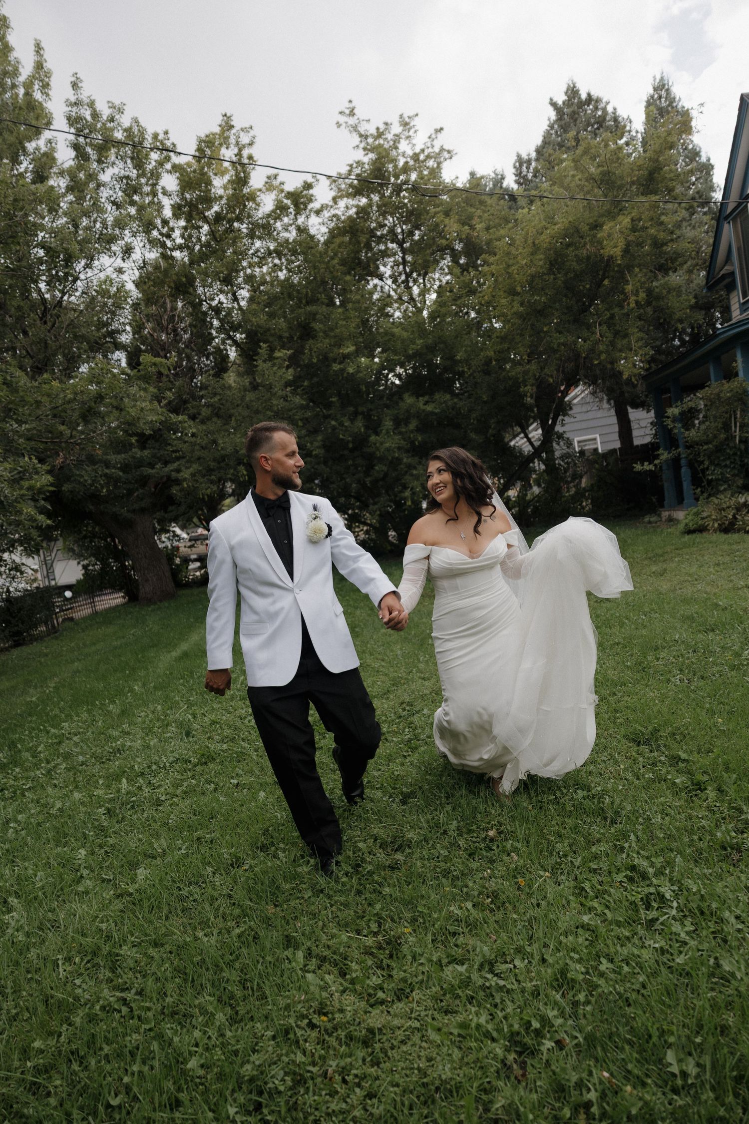 A smiling bride in a white off-shoulder gown and groom in white jacket hold hands walking across green grass.
