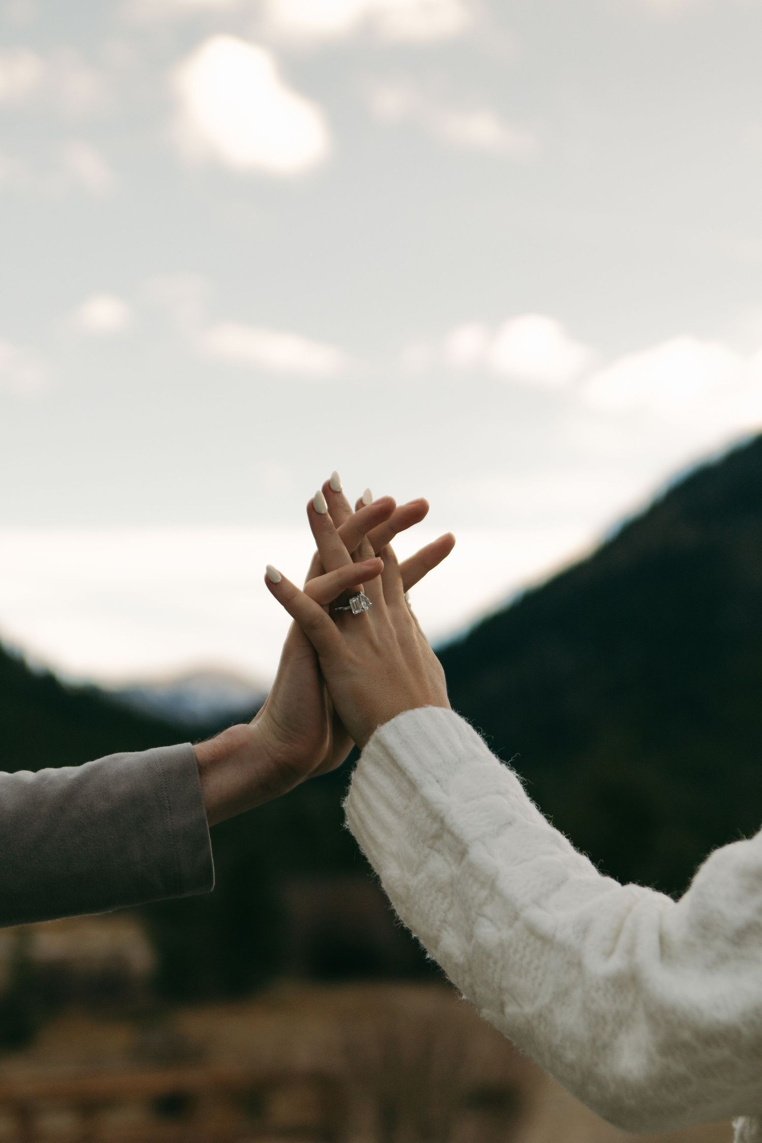 Two hands reach towards each other against a cloudy mountain landscape, with one person wearing a white knit sweater.