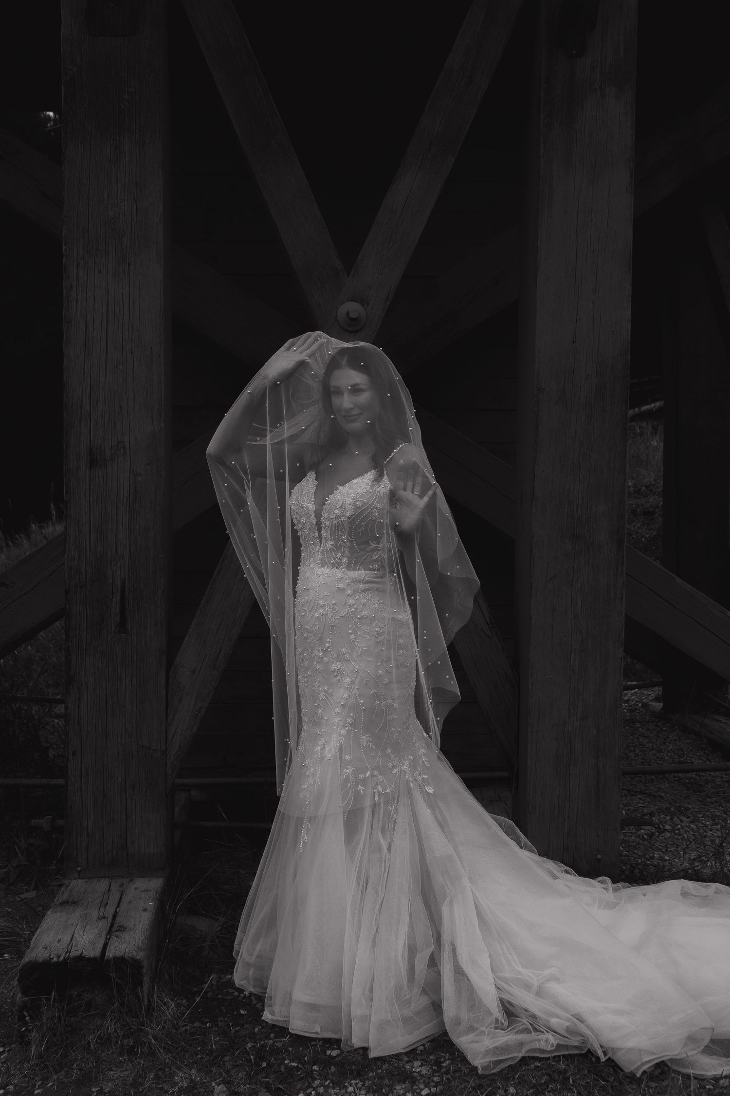 A bride in a flowing lace gown poses under wooden beams in black and white.