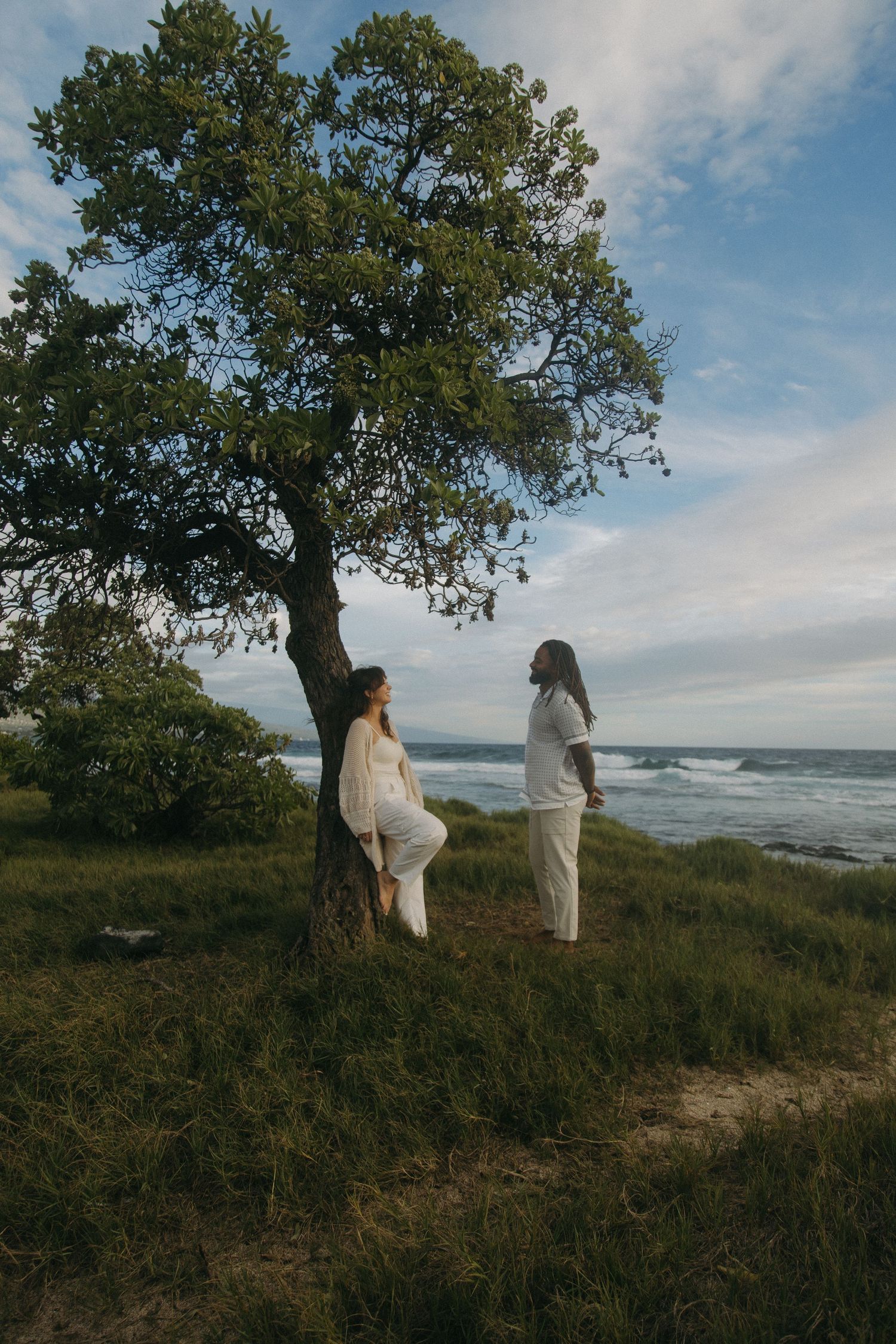 Two figures in white clothing stand near a large tree on a coastal hillside overlooking the ocean on a cloudy day.