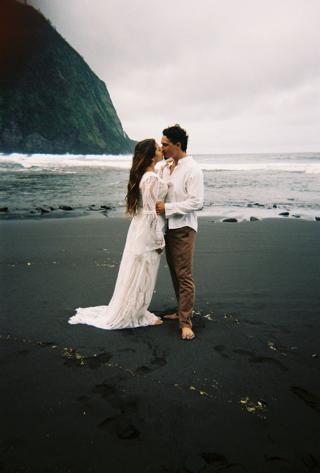 A romantic couple in wedding attire shares an intimate moment on a dramatic black sand beach with cliffs and ocean waves.