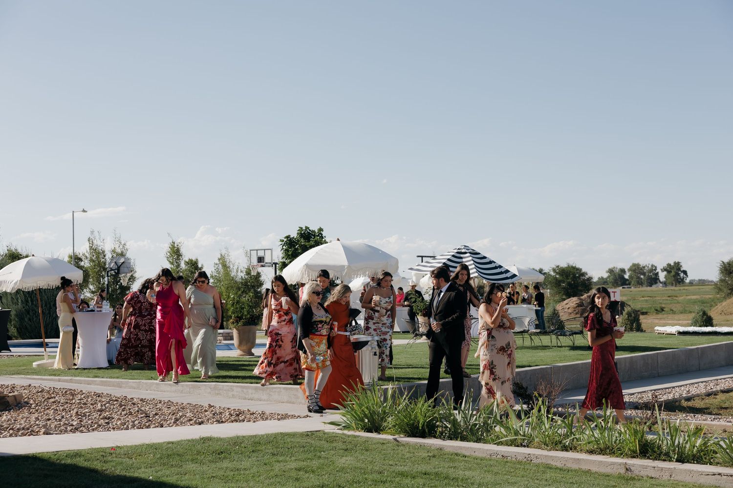 Guests in colorful traditional attire walk along a garden pathway at an outdoor wedding ceremony on a sunny day.