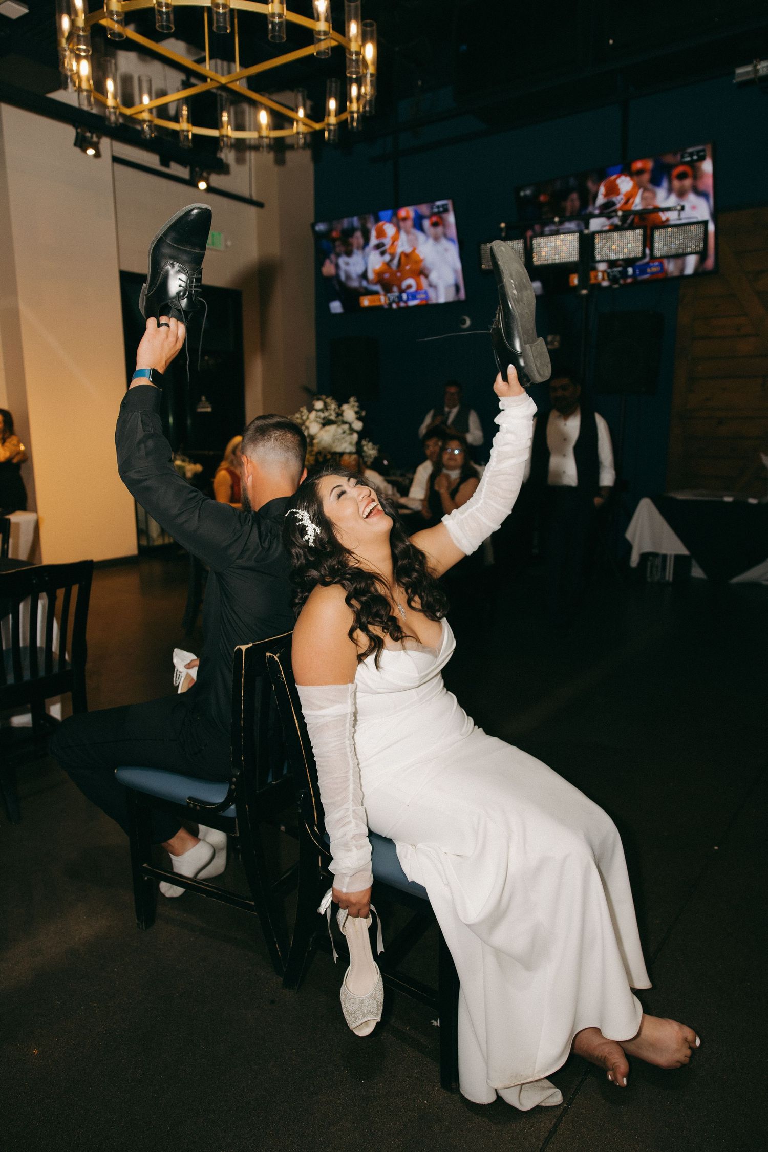 Bride and groom playing shoe game at wedding reception raising footwear under gold chandelier.