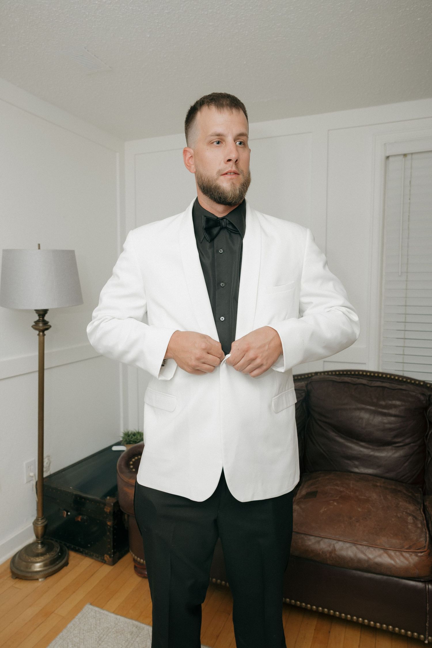 Groom in white blazer and black shirt buttoning jacket indoors before wedding ceremony.