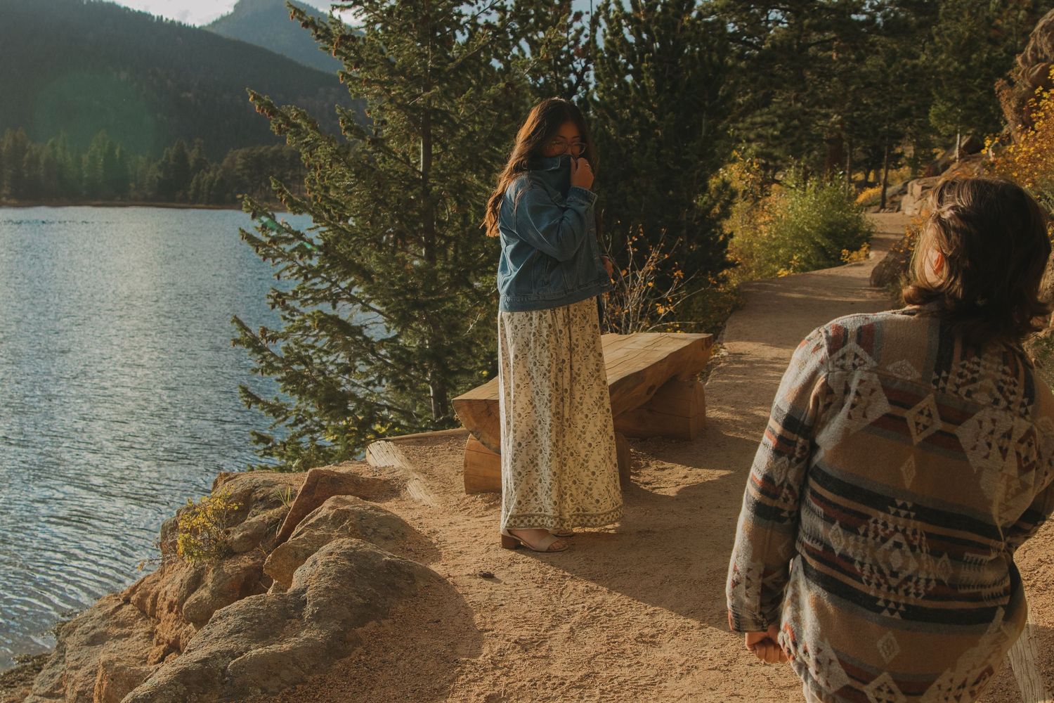 A scenic mountain trail winds along a tranquil lake shore with evergreen trees and rocky outcrops in the background.