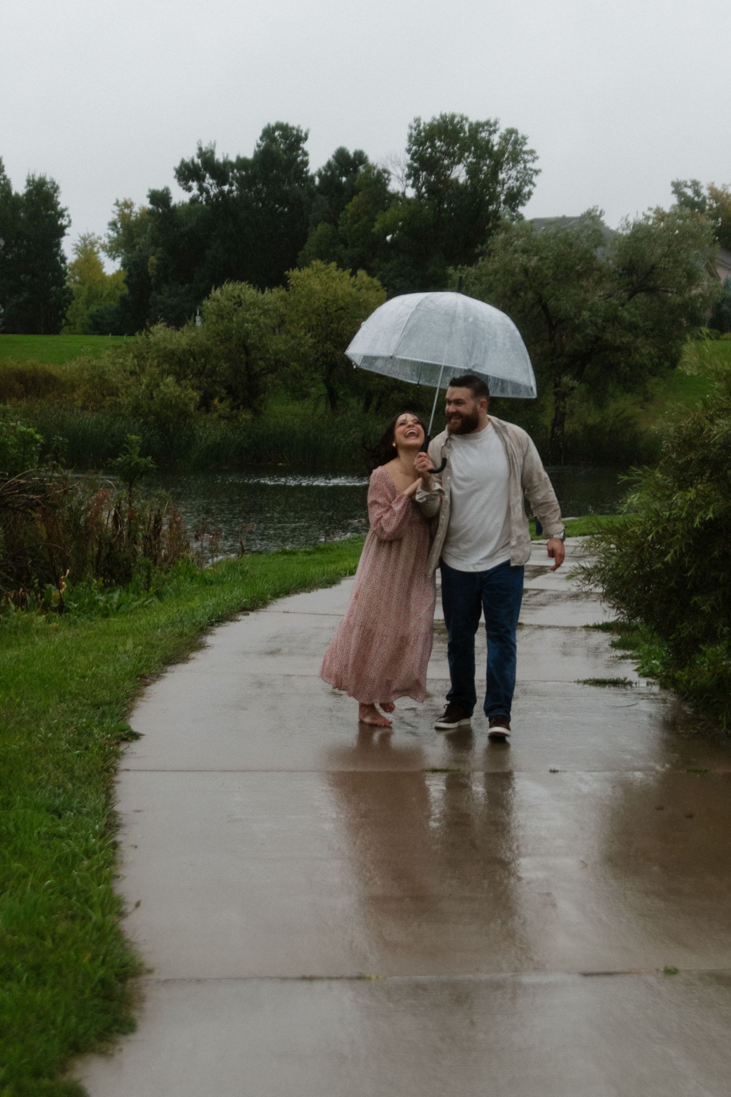 A couple shares a romantic walk in the rain under a clear umbrella on a wet pathway surrounded by greenery.
