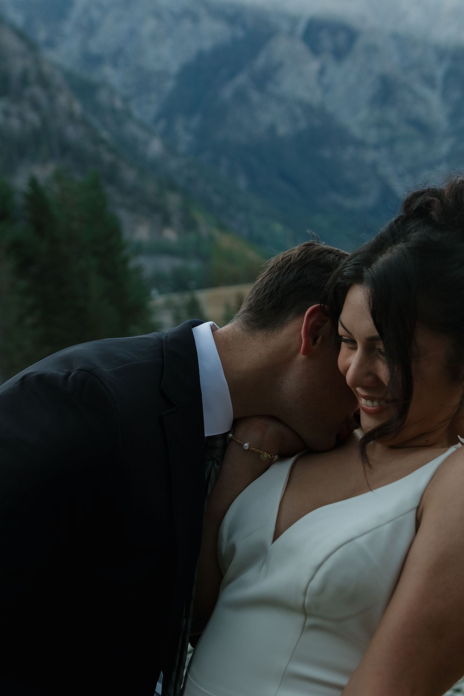 A romantic wedding moment captured against a mountain backdrop, showing a sweet gesture between two people.