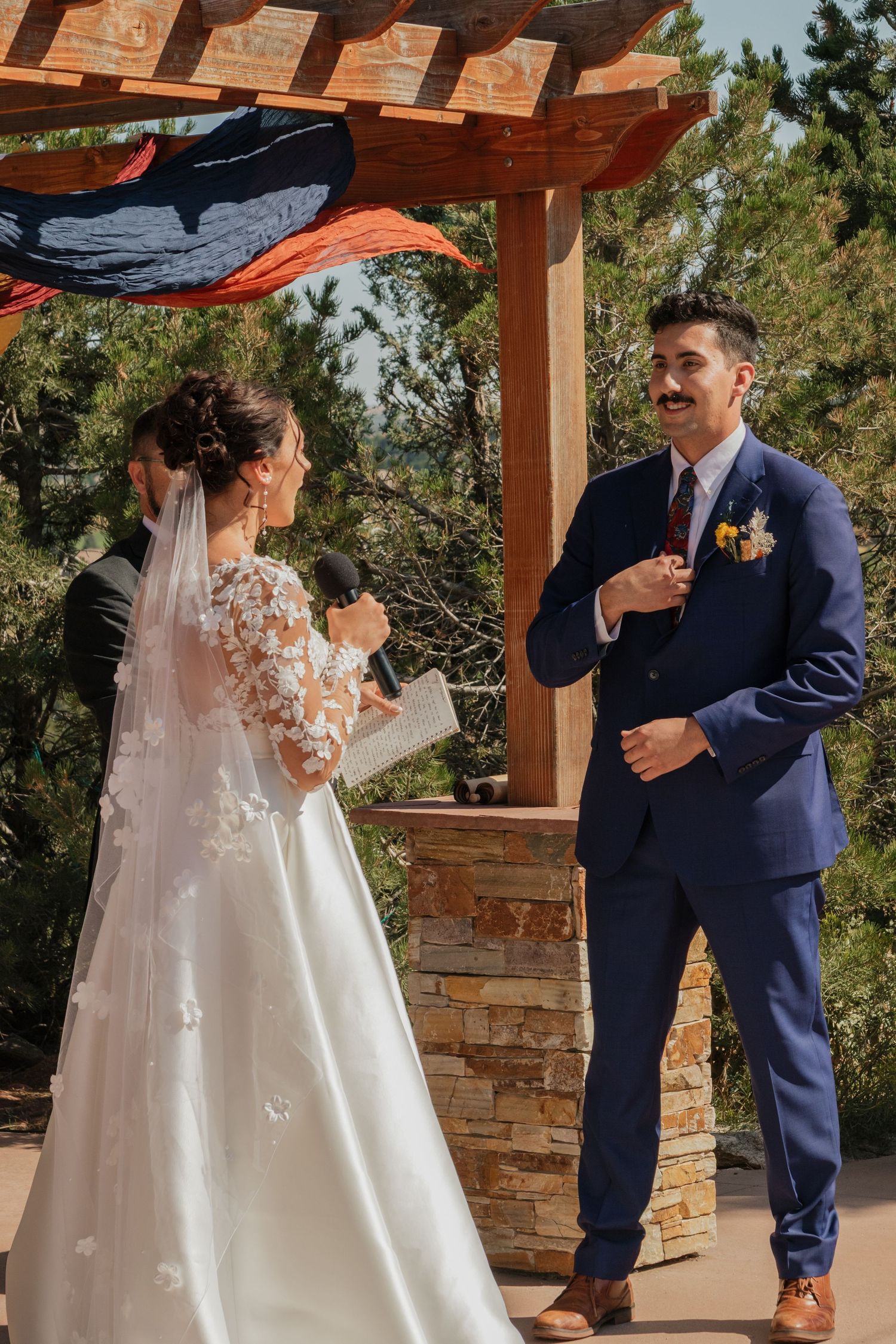 Couple exchanges vows during outdoor ceremony under rustic wooden pergola with scenic mountain views.