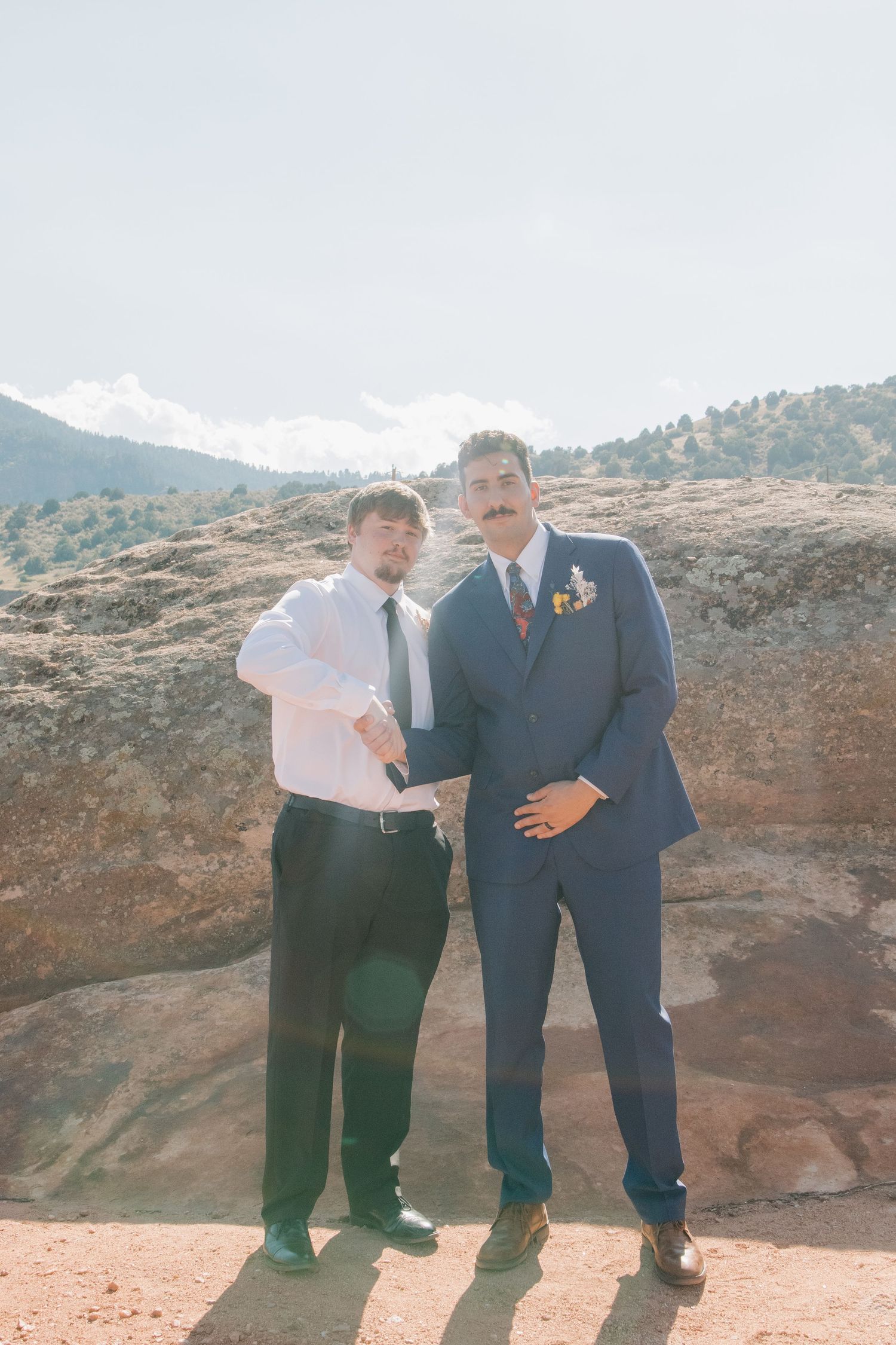 Two wedding attendees pose together against a mountain backdrop on a sunny day.