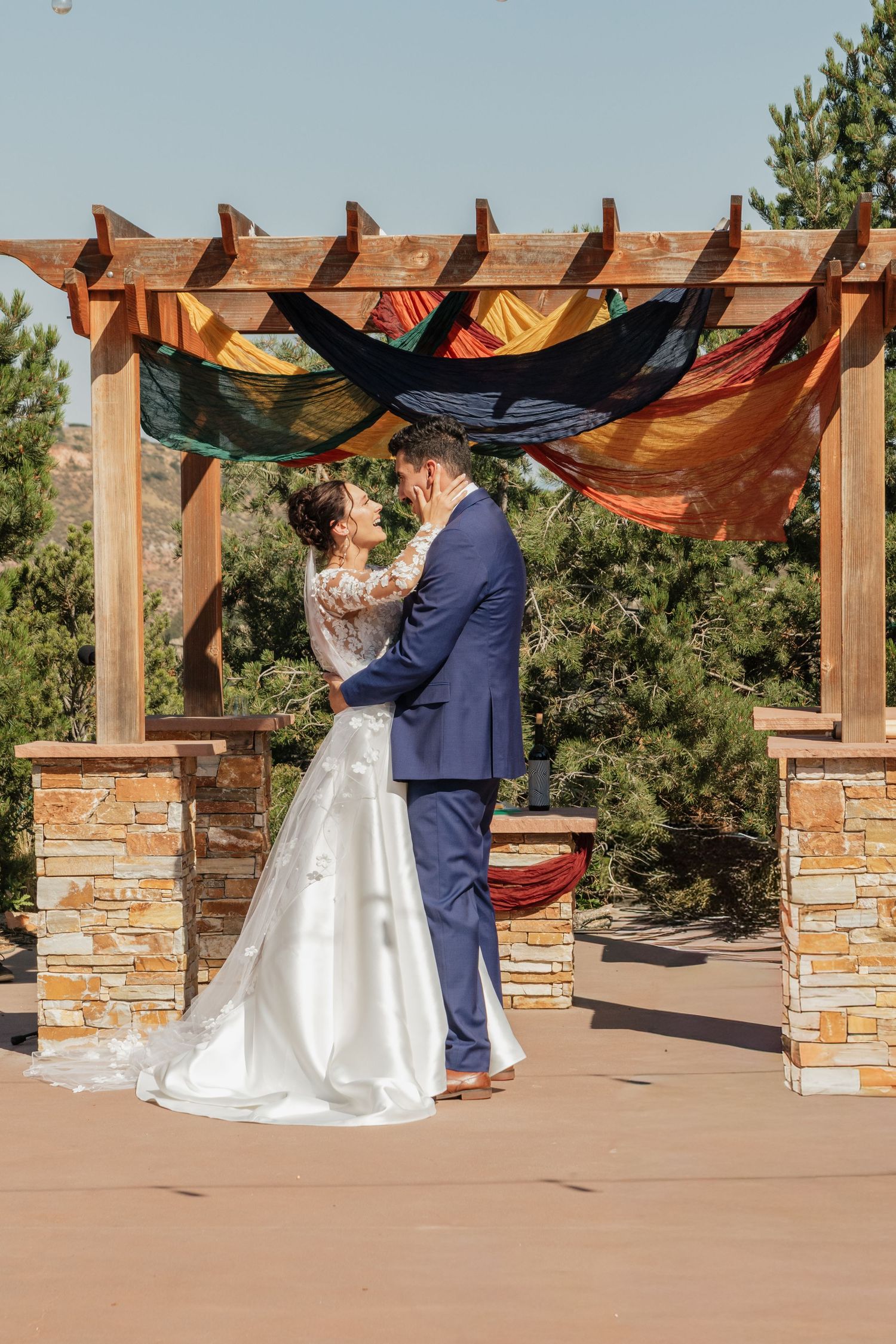 Wedding couple embracing under a rustic wooden pergola with stone pillars and mountain scenery.