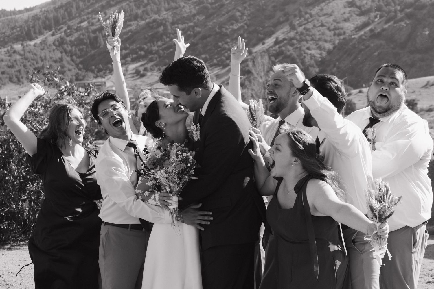 Wedding party jumping and celebrating with excitement outdoors against mountain scenery.