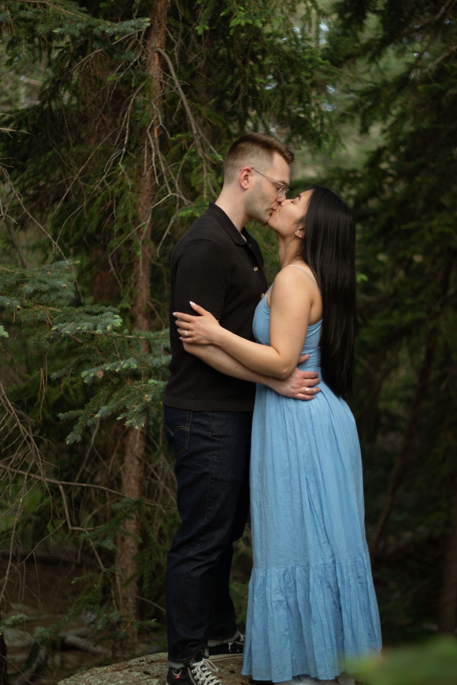 A couple sharing an intimate moment among evergreen trees in casual attire.