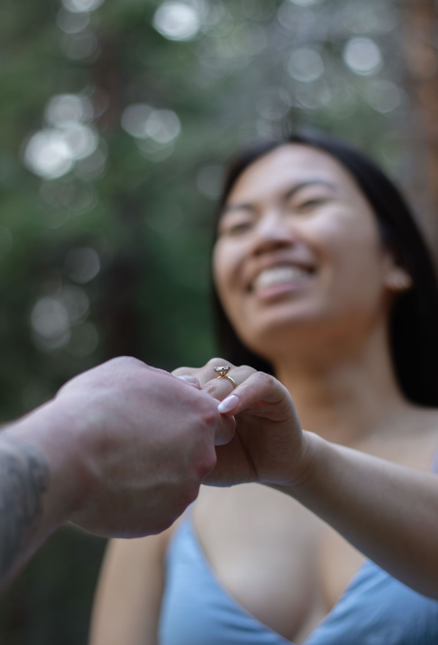 A heartfelt moment captures a close-up of hands with an engagement ring against a bokeh forest background.