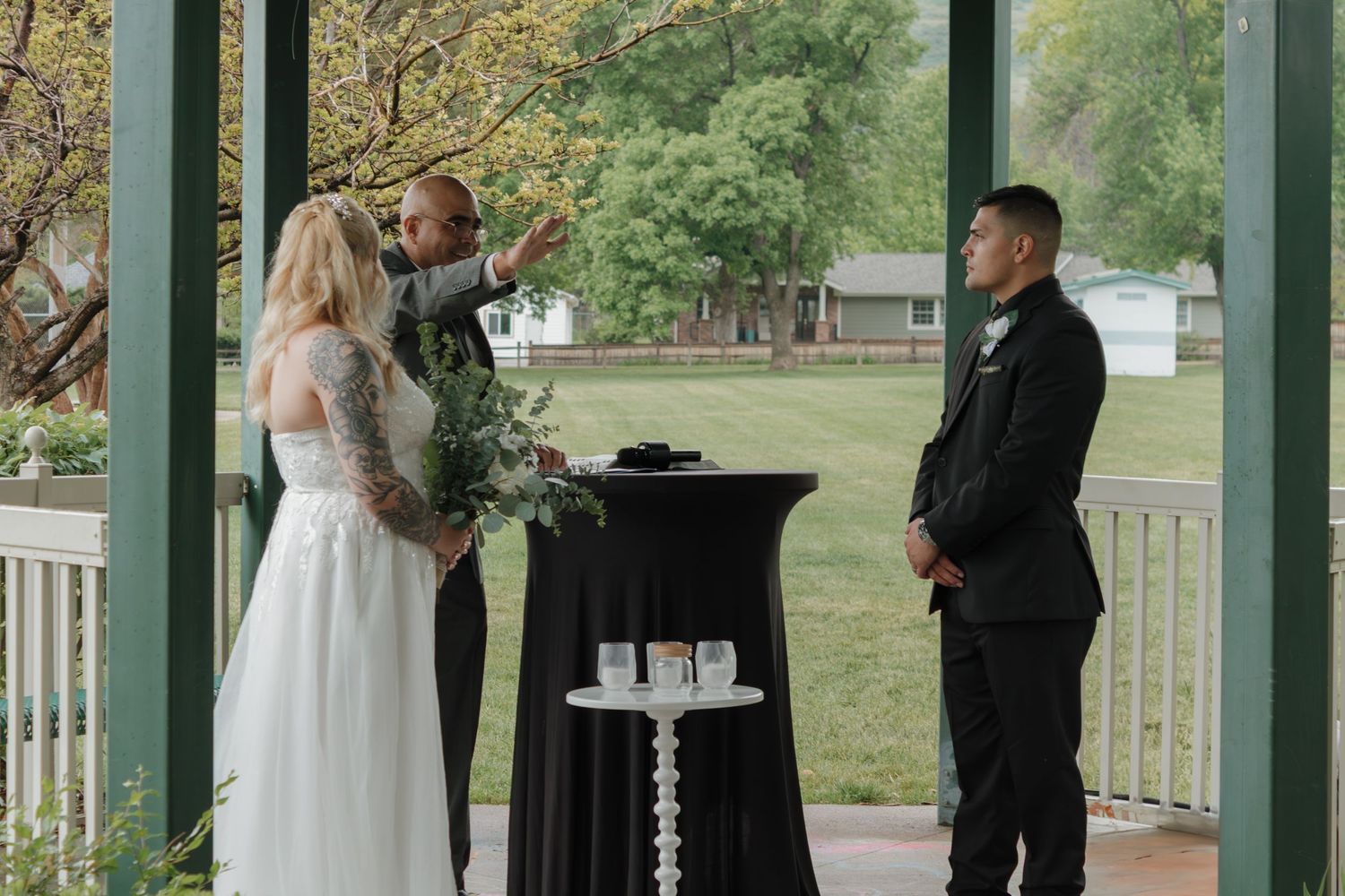 An intimate wedding ceremony unfolds in a park gazebo with lush greenery in the background.