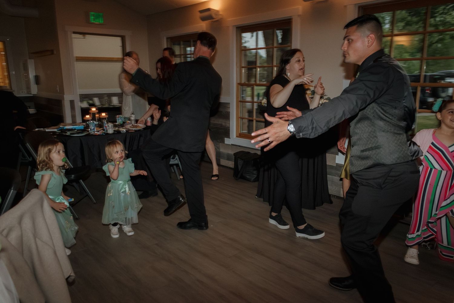 People dancing and celebrating at a wedding reception with children in formal attire on a hardwood floor.