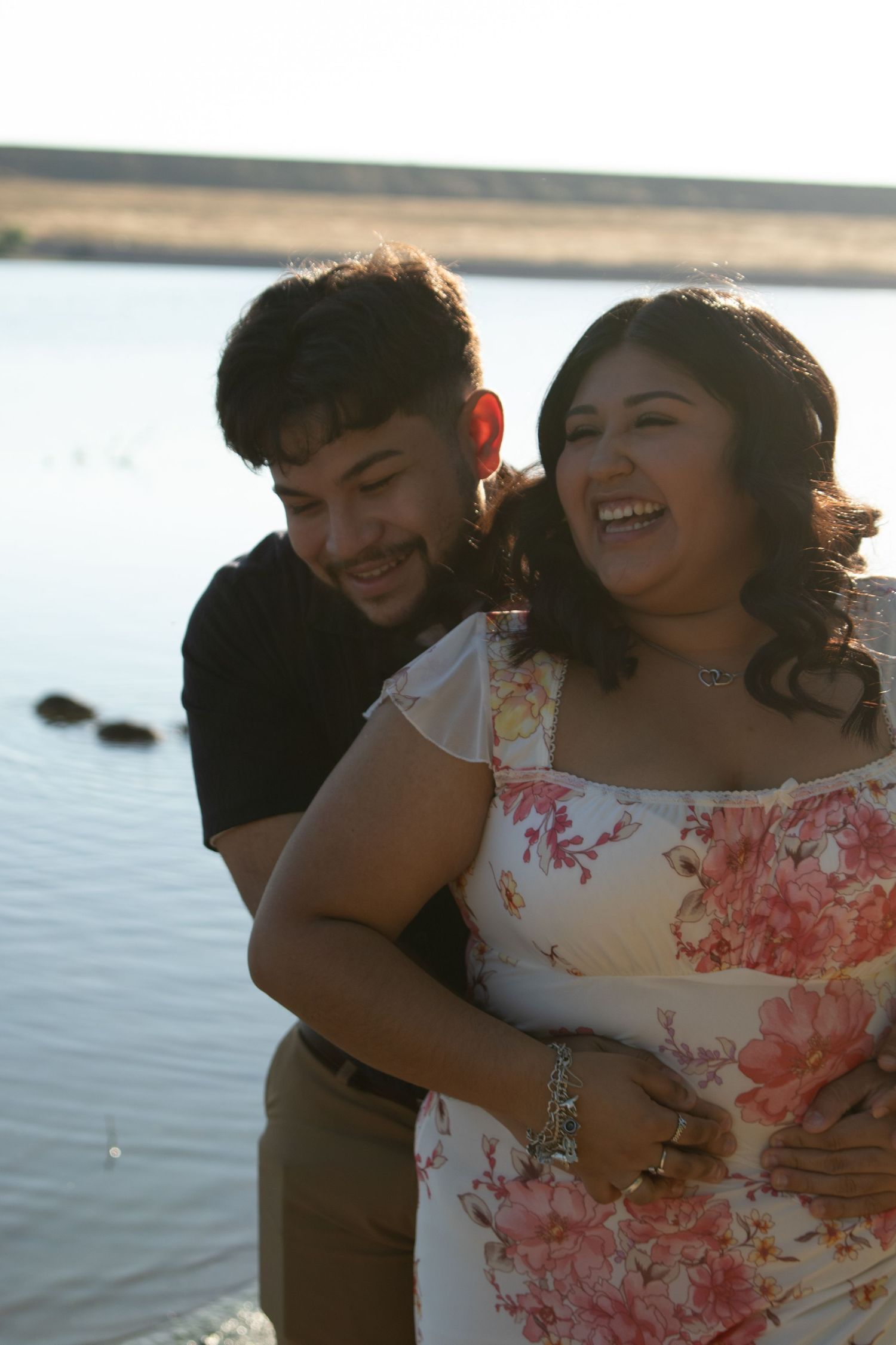A couple shares a tender embrace at sunset by a calm waterfront, with warm natural lighting creating a romantic atmosphere.