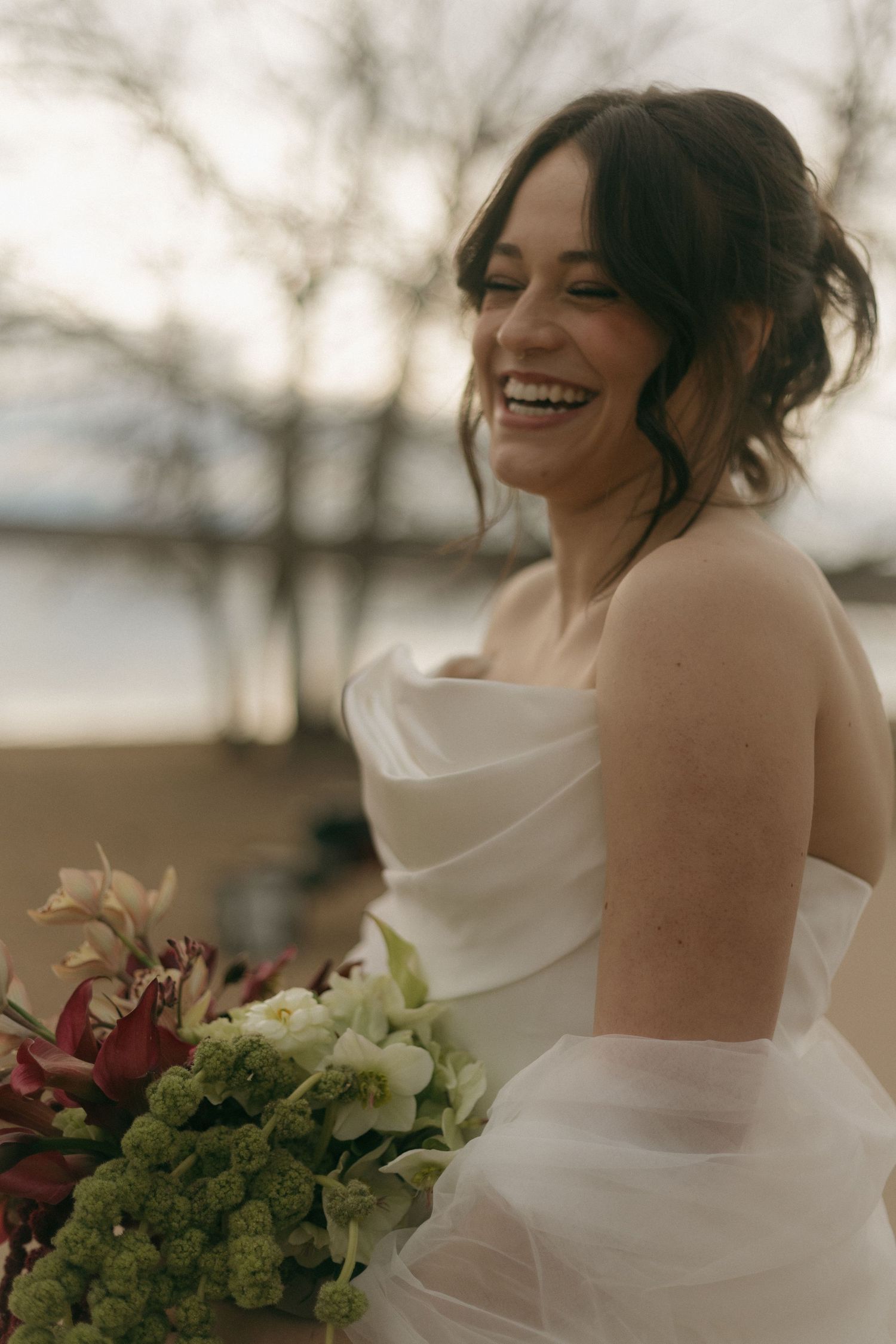 Bride in white strapless gown holding burgundy and green bouquet laughs joyfully during outdoor wedding photoshoot.