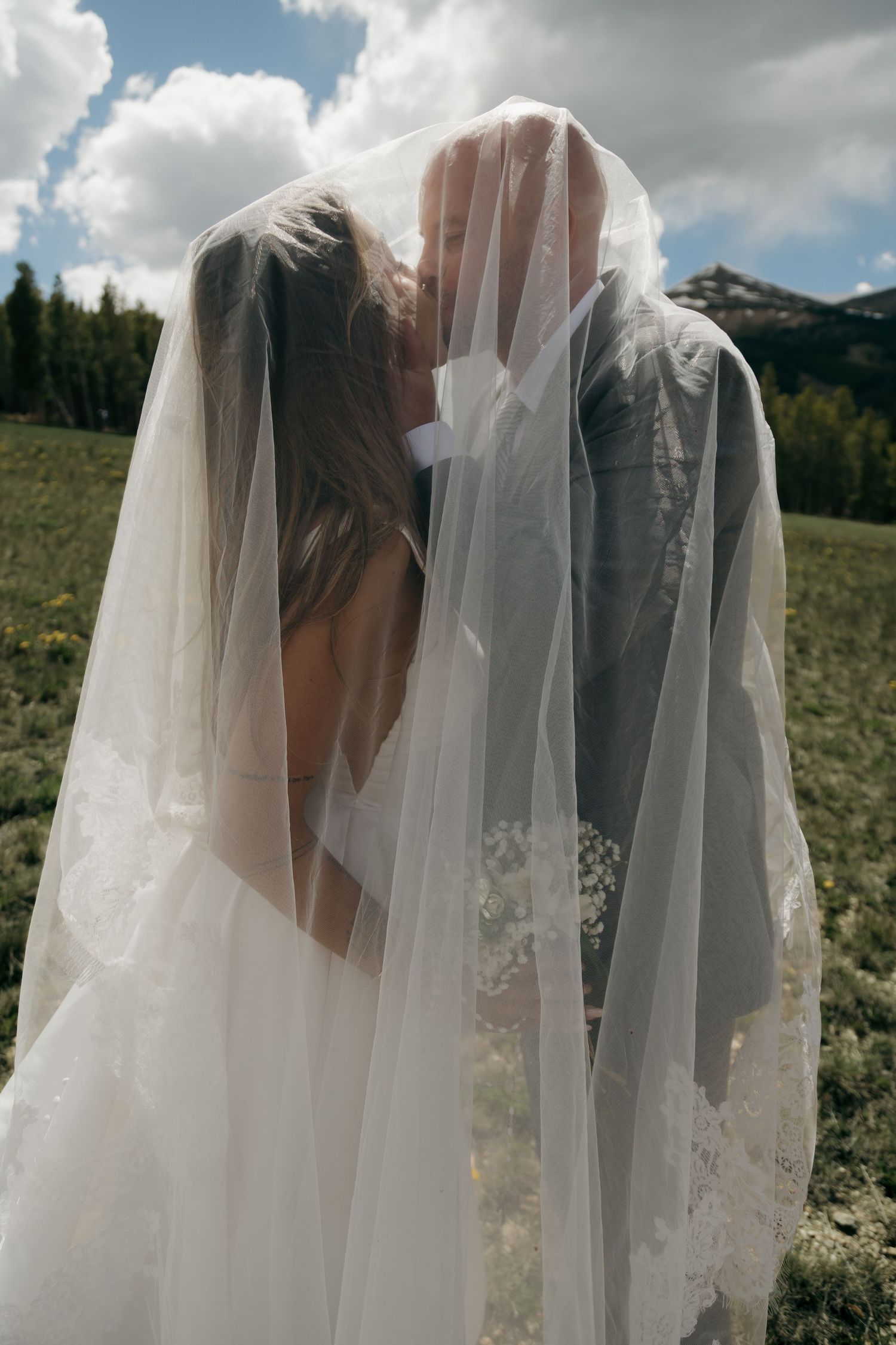 Bride and groom sharing a kiss hidden beneath a flowing wedding veil in a mountain meadow.