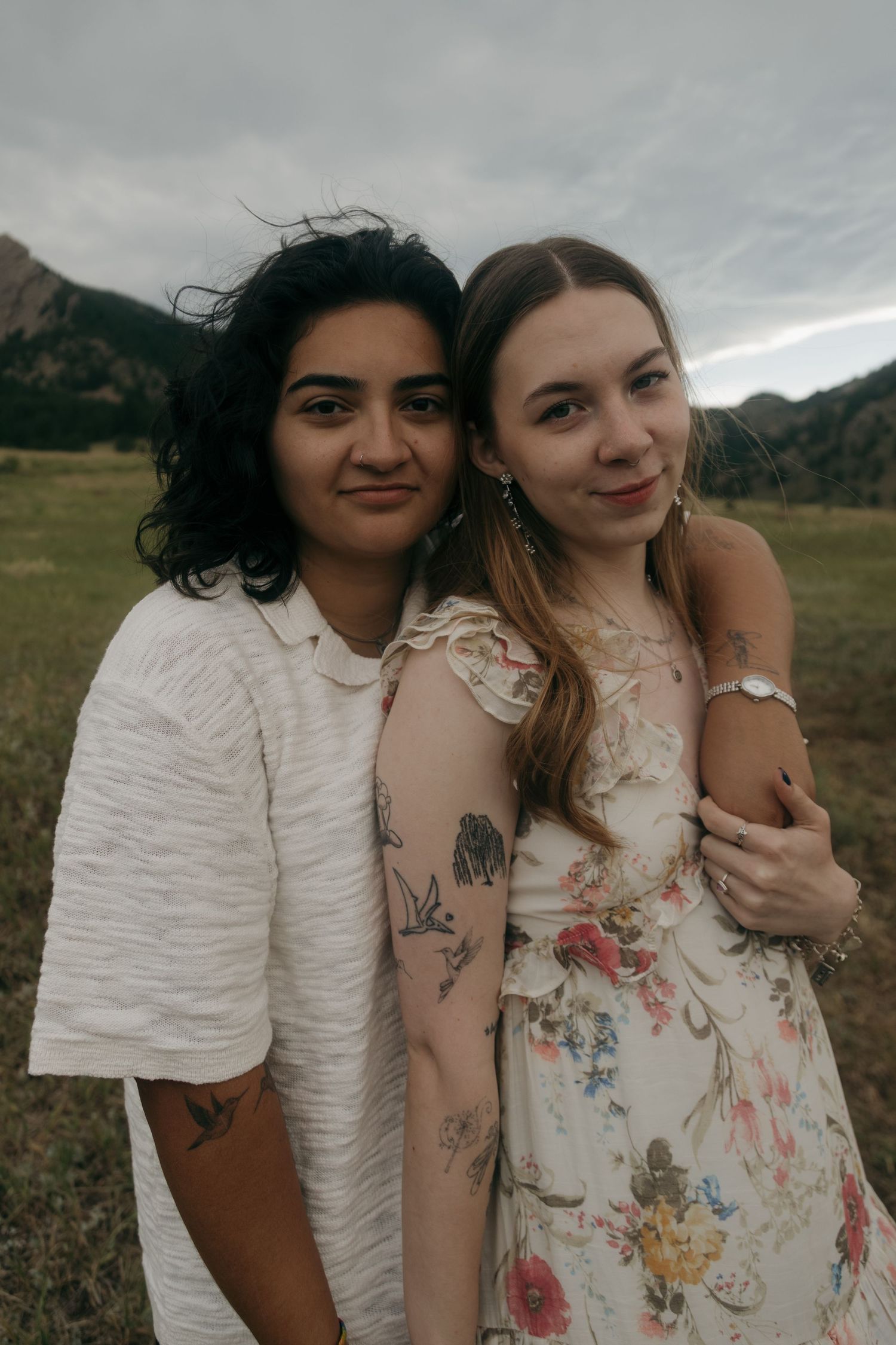 Two women embracing outdoors in a field with mountains, one in white textured top and one in floral dress with tattoos.