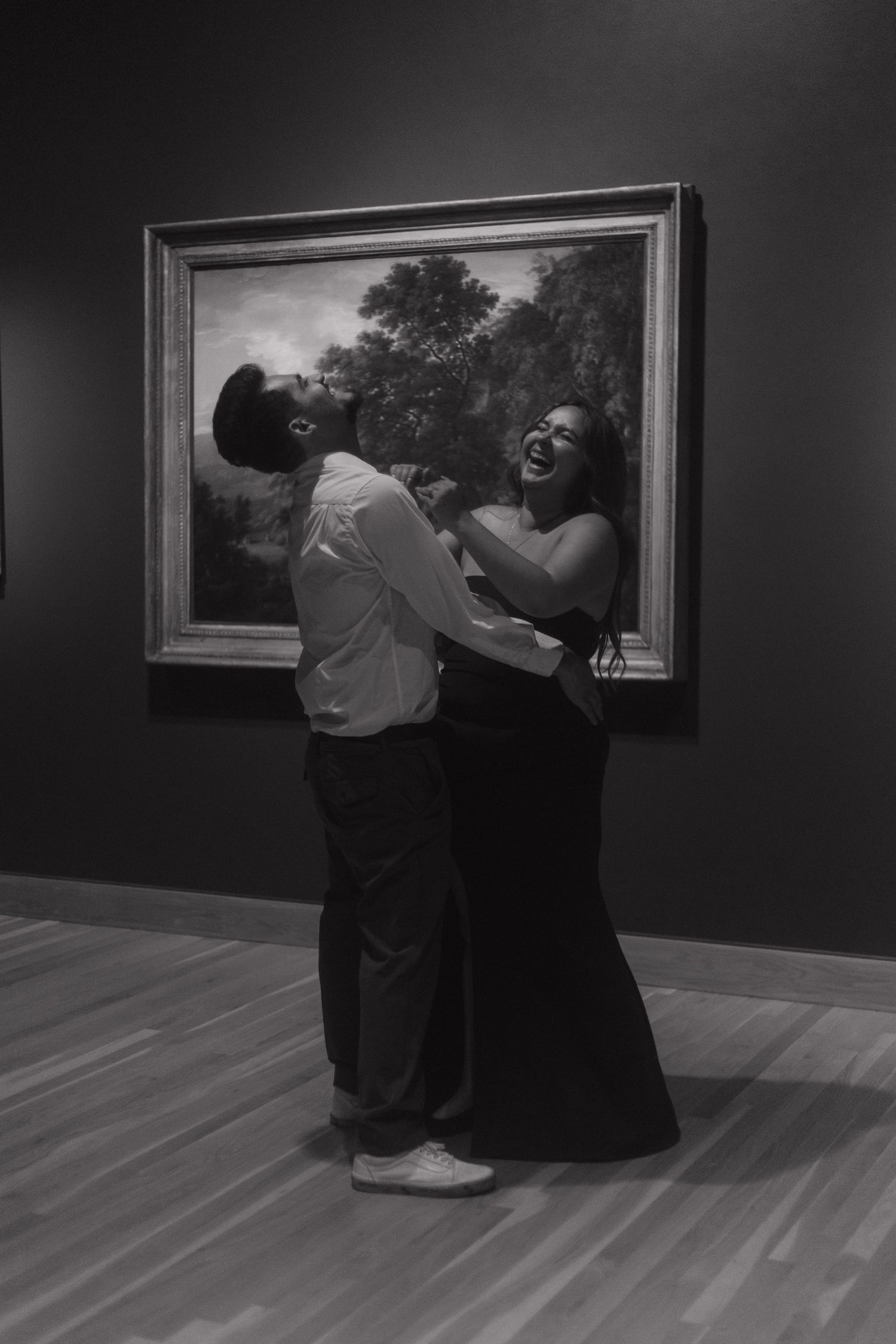 Black and white photo of a laughing couple dancing playfully in front of a classic painting.