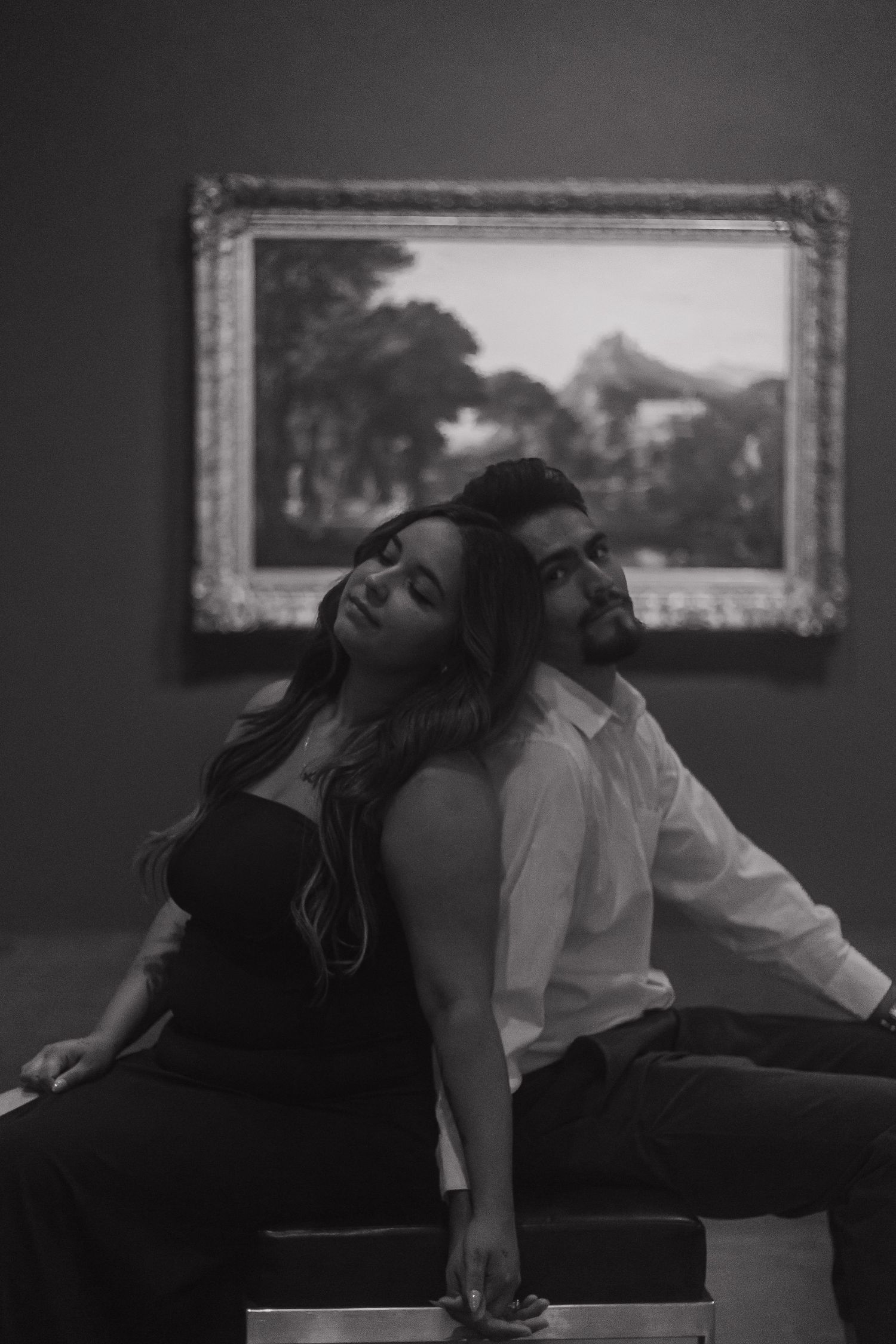 Black and white photo of a couple sitting back-to-back in a museum gallery with a landscape painting behind them.