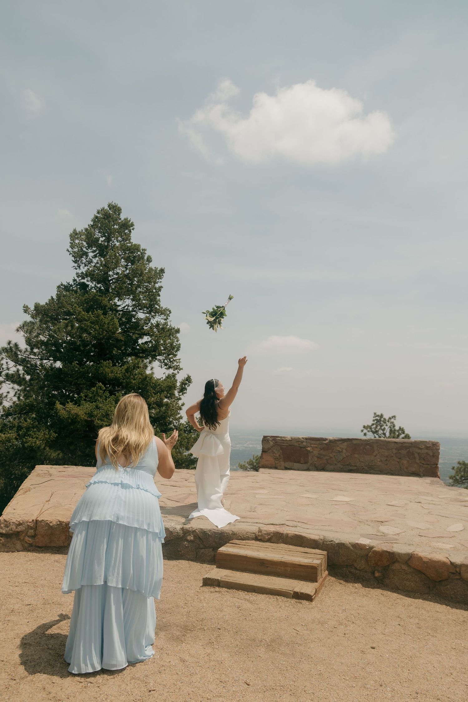 A bride tosses her bouquet into the air on a stone terrace while a bridesmaid in blue watches nearby.