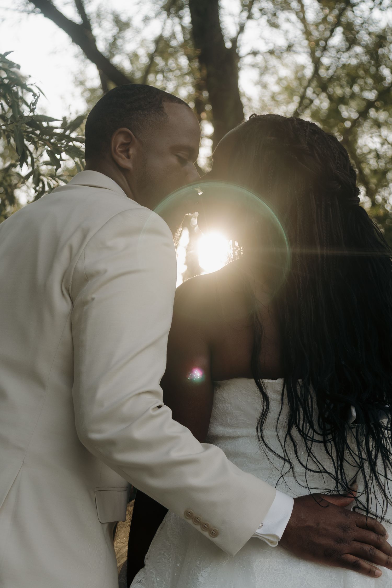 Black couple sharing romantic kiss with golden sun flare, groom in white suit embracing bride in wedding dress outdoors.
