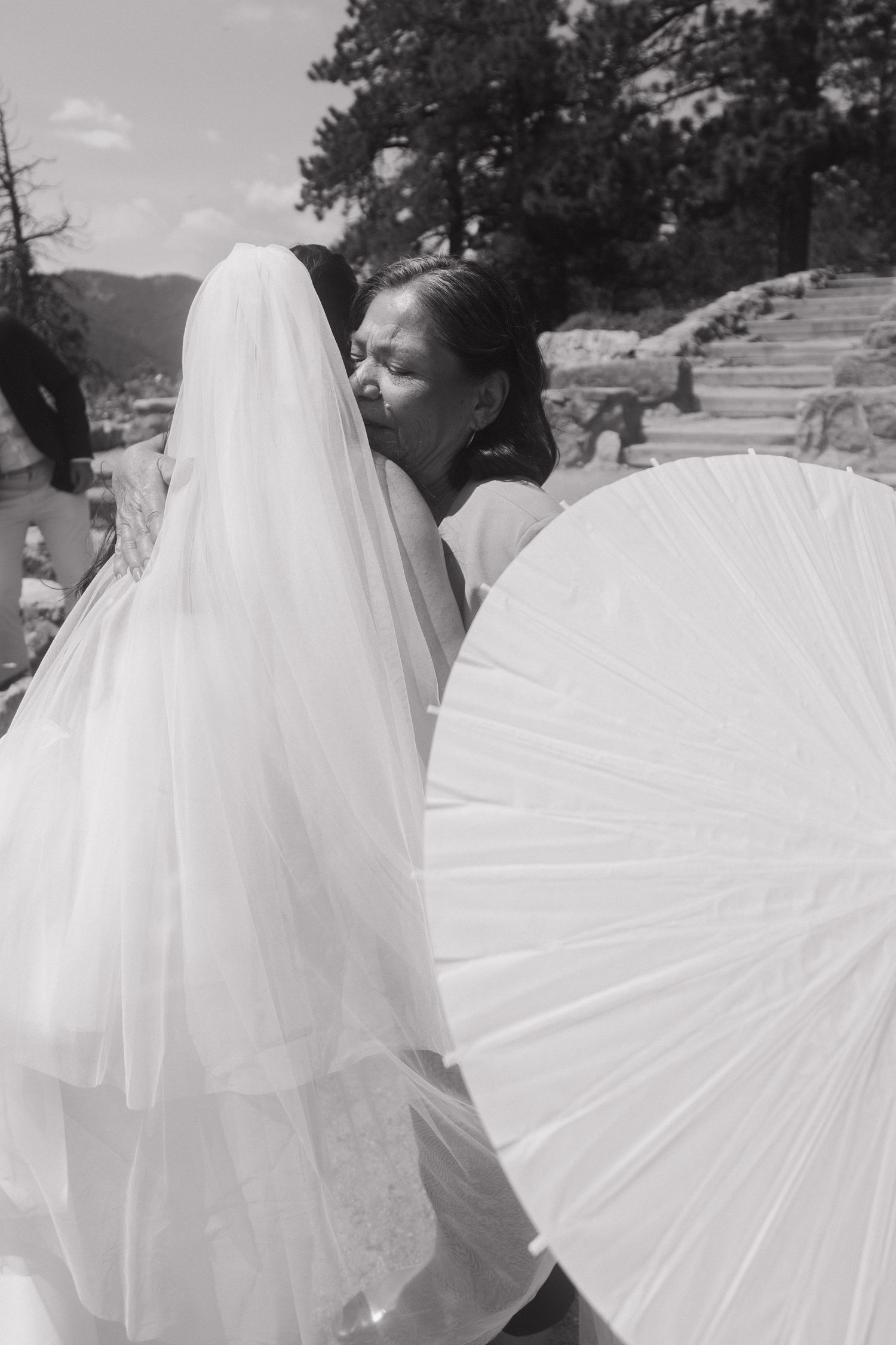 Bride in white veil and gown embraces guest while holding parasol at outdoor wedding ceremony.