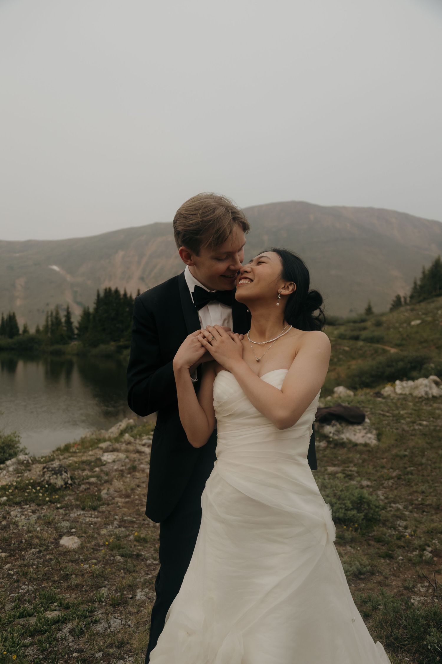 Groom tenderly nuzzles bride as she smiles, both holding hands near a serene alpine lake with mountain backdrop.