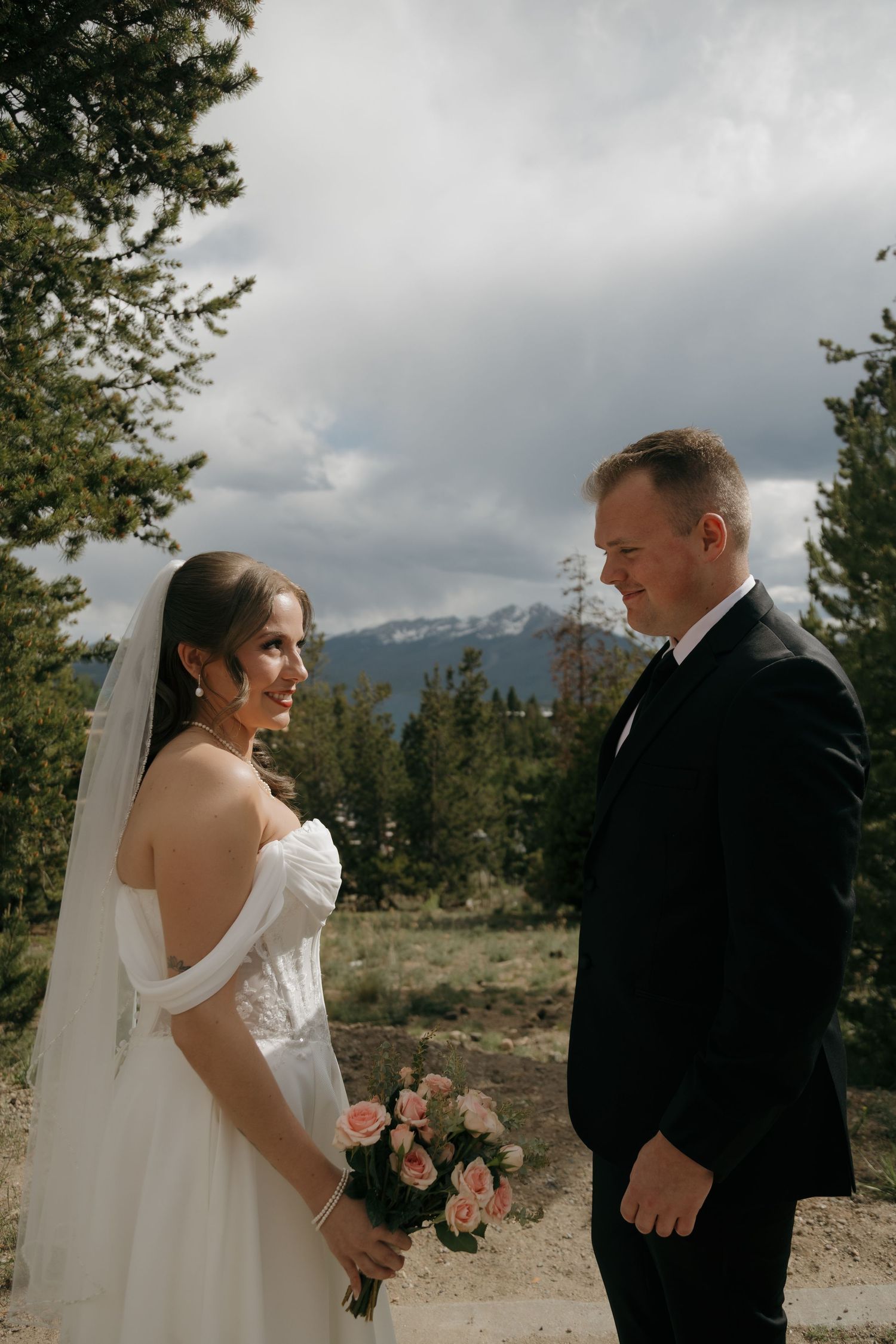 Bride holding pink bouquet and groom share a tender moment with snow-capped mountains in the background.