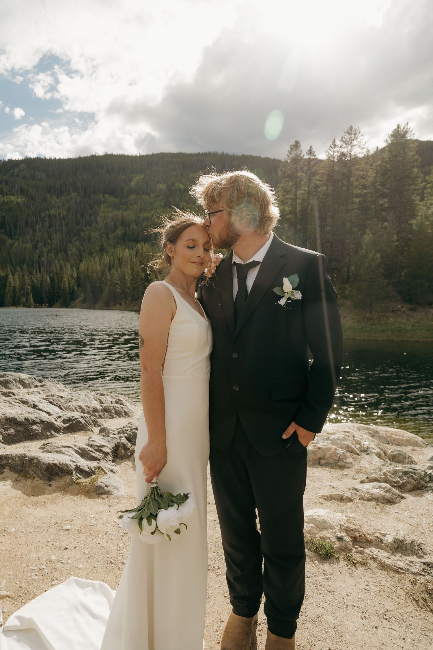 Bride and groom embrace romantically by sunlit mountain lake, groom kissing bride on cheek.