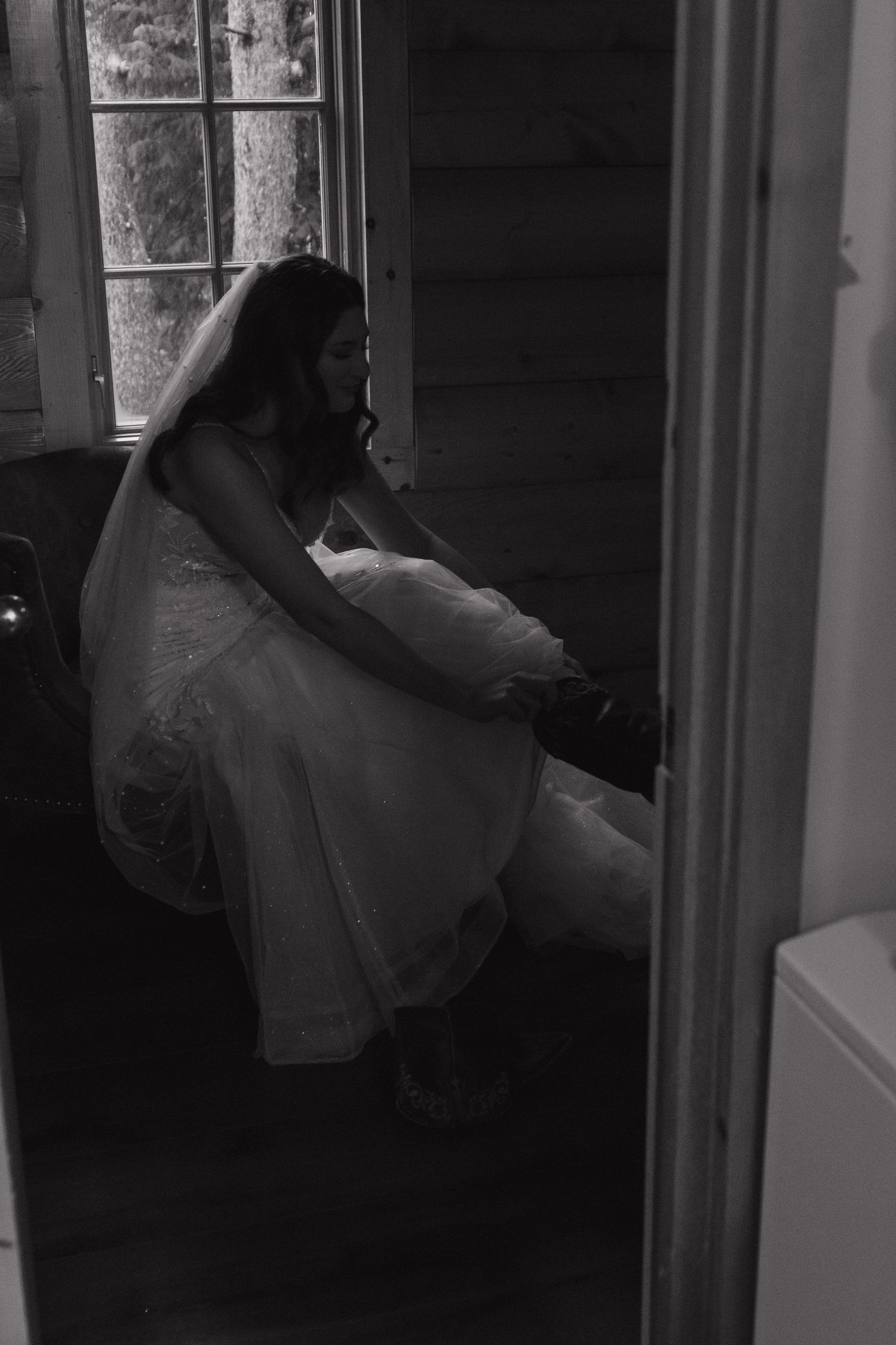 A moody black and white photo of a bride in a wedding dress sitting alone in a dark room.