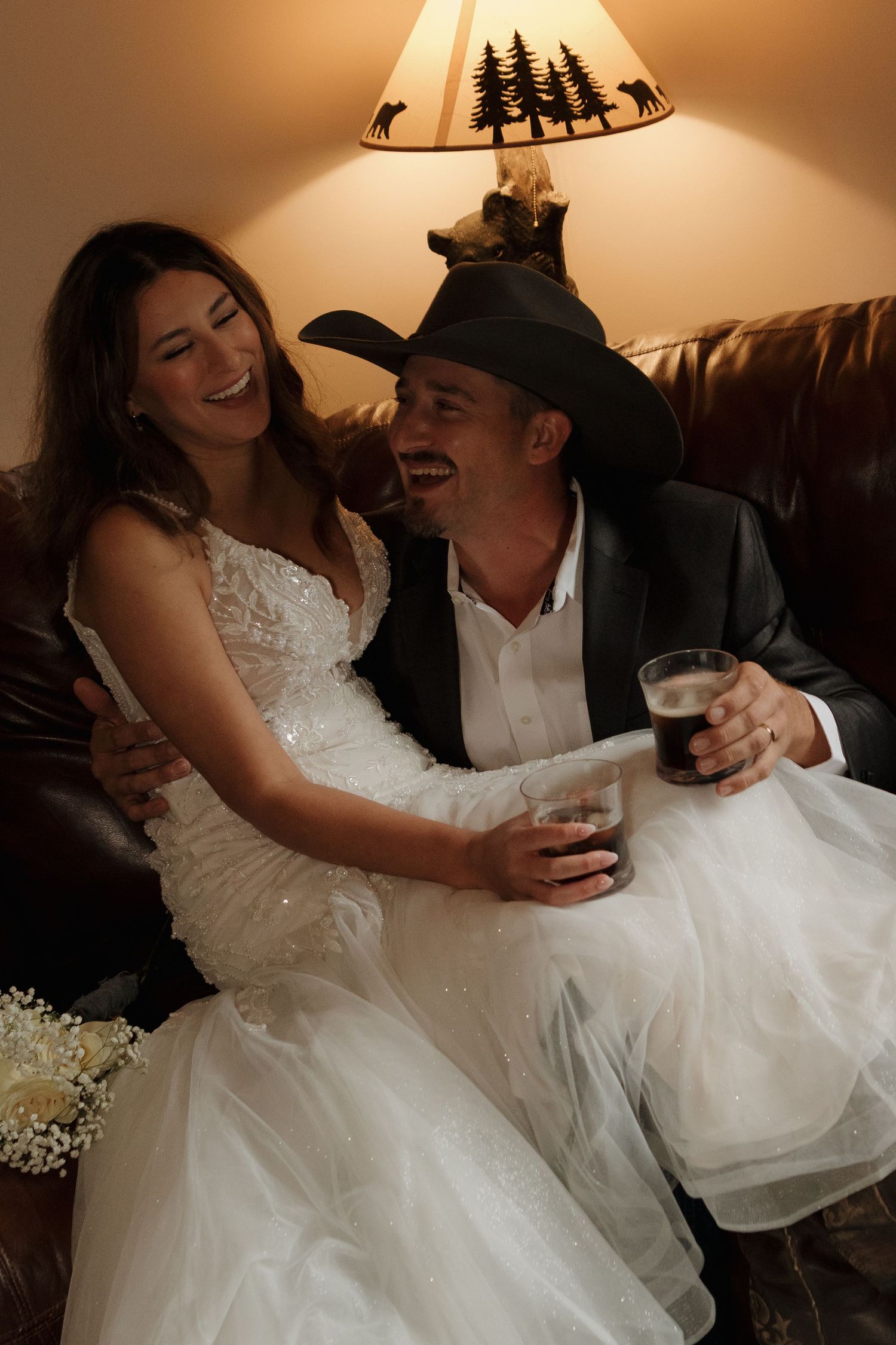 A joyful wedding couple shares drinks and laughter while sitting on a couch under a rustic lamp.
