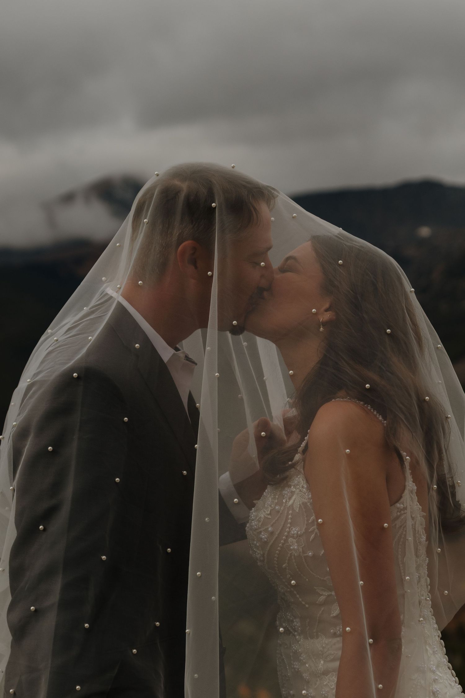 A romantic wedding couple shares an intimate kiss under a delicate veil against a moody mountain backdrop.