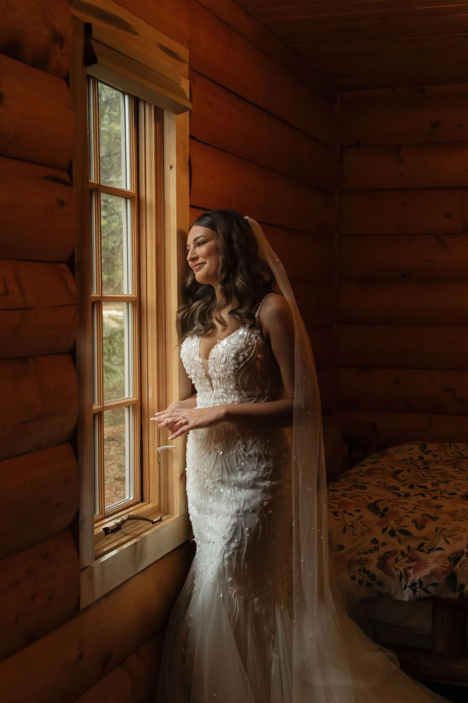 A bride in a lace wedding dress stands by a rustic cabin window in warm natural light.