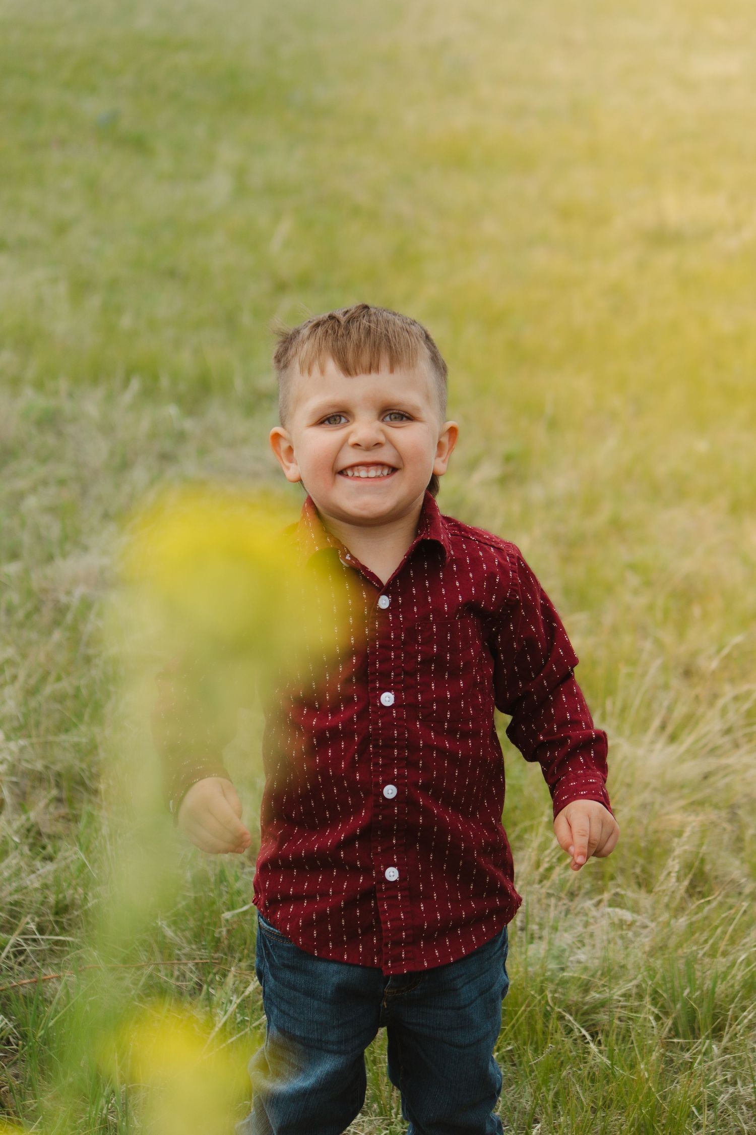 A child wearing a red patterned button-down shirt stands in a sunlit field with tall grass.