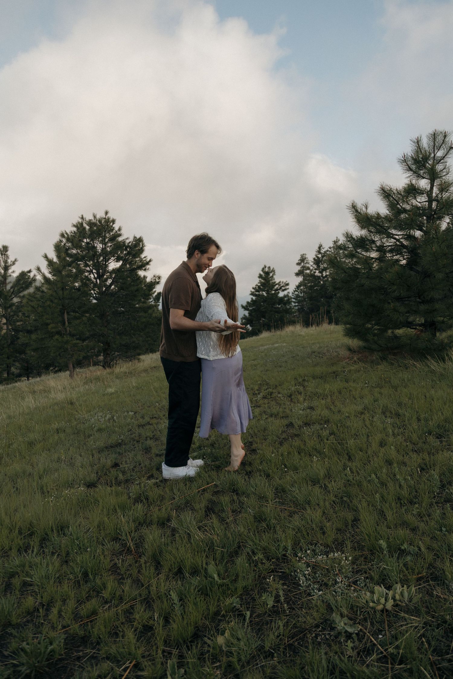 Couple sharing a romantic kiss on a lush green hillside surrounded by pine trees under a cloudy sky.