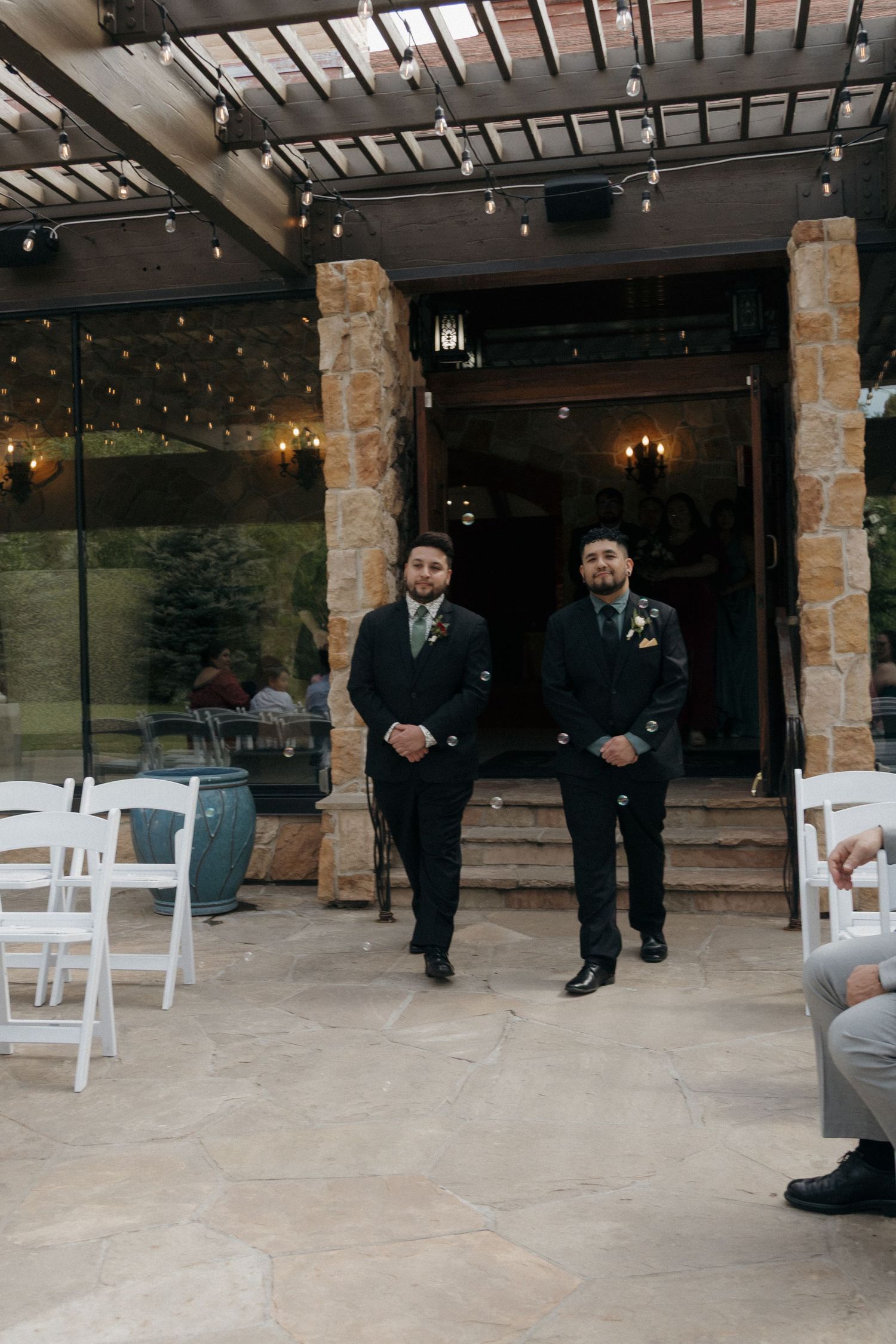 Two groomsmen in black suits walking down the aisle at an outdoor wedding ceremony with string lights.
