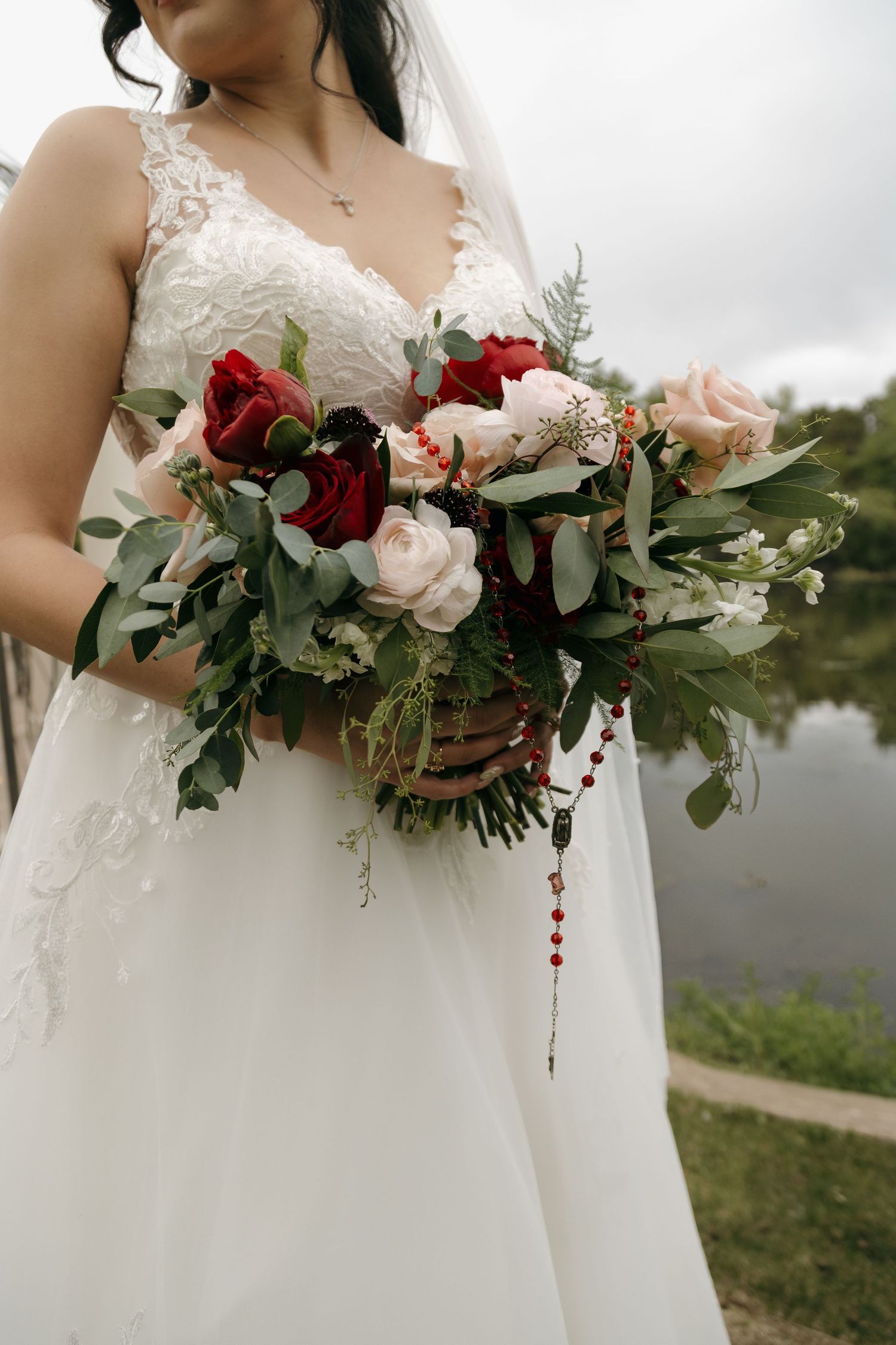 Bride holding a lush wedding bouquet with red and blush roses, greenery, and a rosary, wearing a white lace gown.
