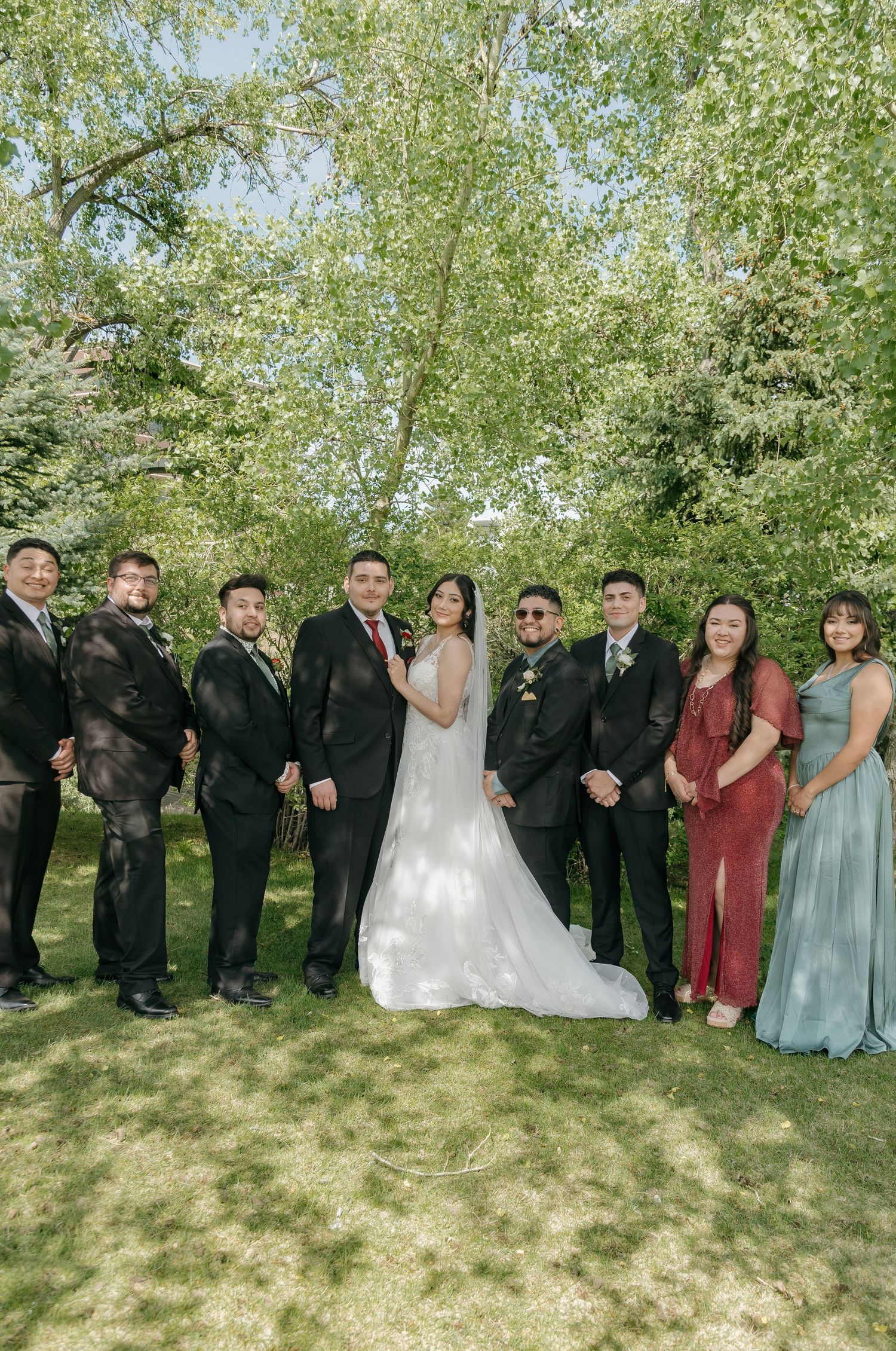 Wedding party poses outdoors in lush green garden, groomsmen in black suits flanking bride in white gown and groom.