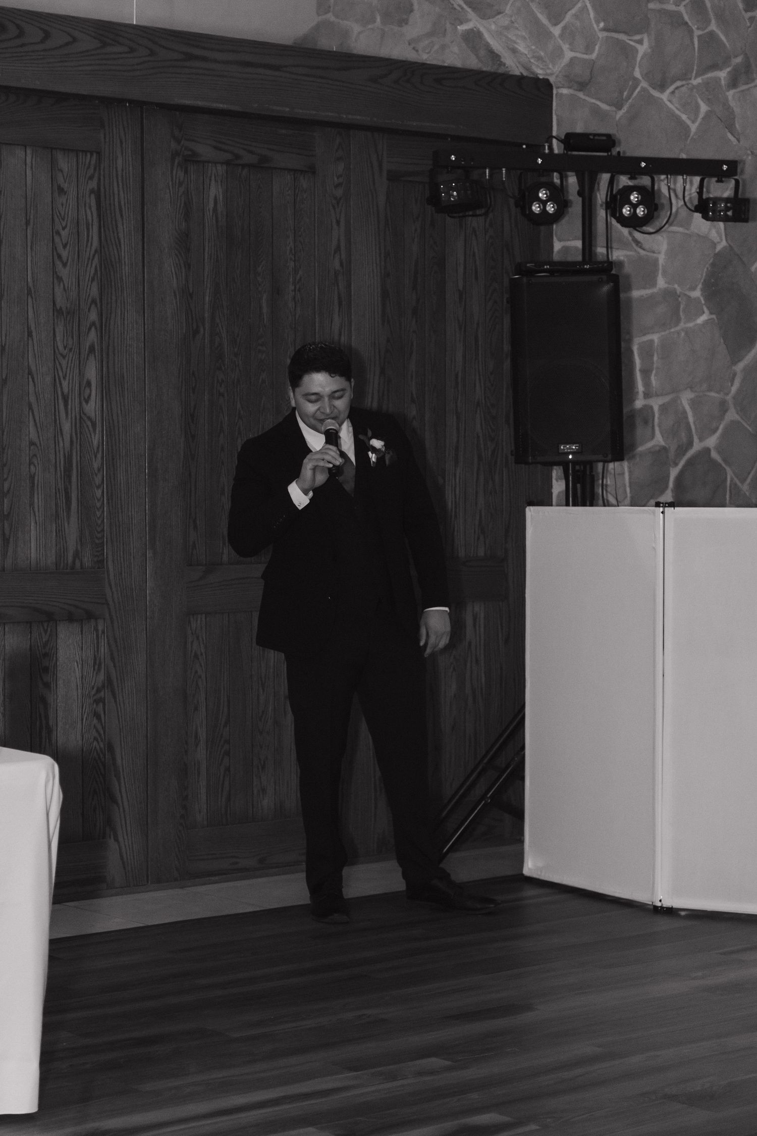 Black and white photo of groom in tuxedo speaking into microphone during wedding reception toast near DJ setup.
