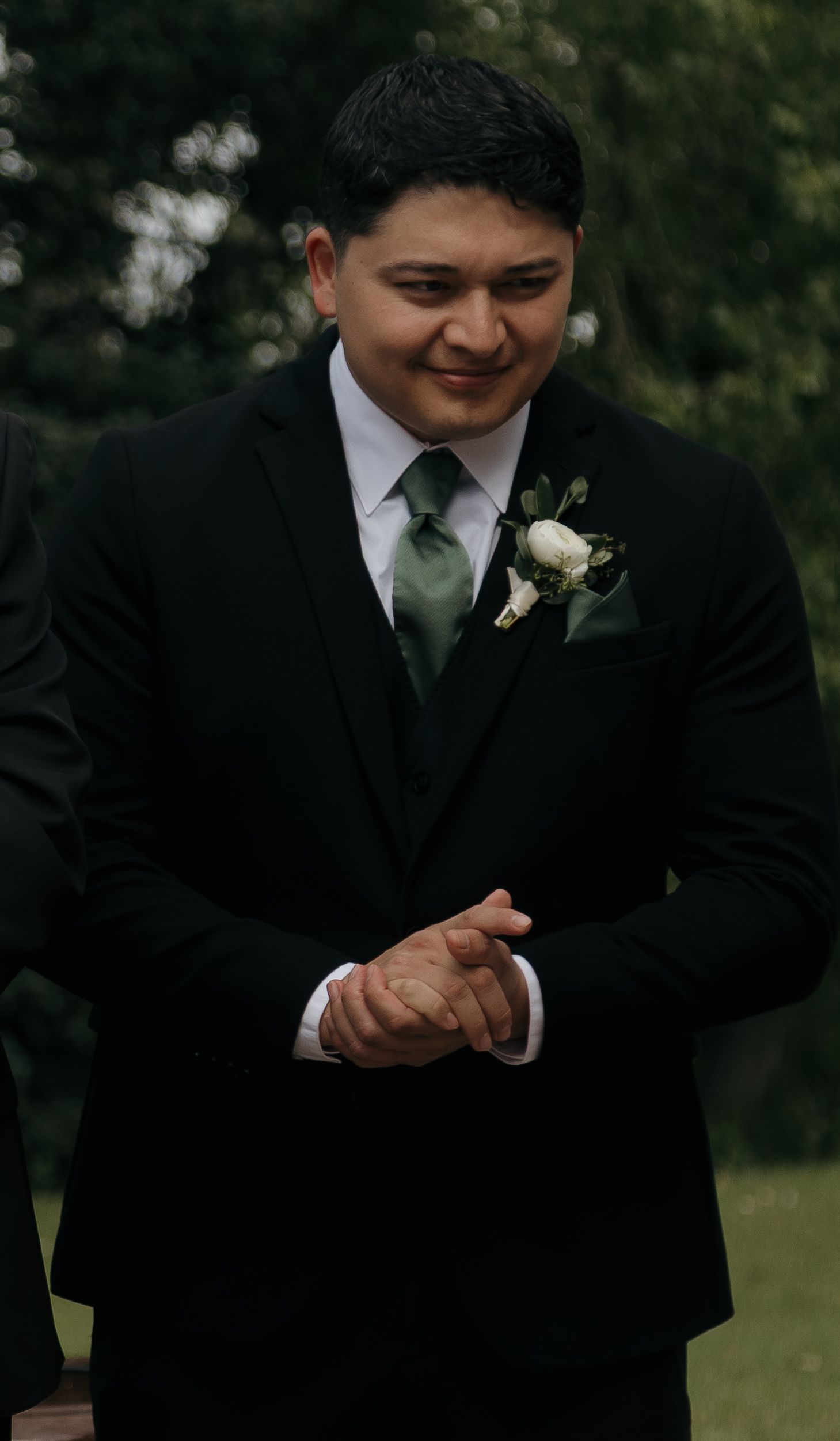 Smiling groom in black suit with white boutonniere clasps his hands while waiting at the ceremony.