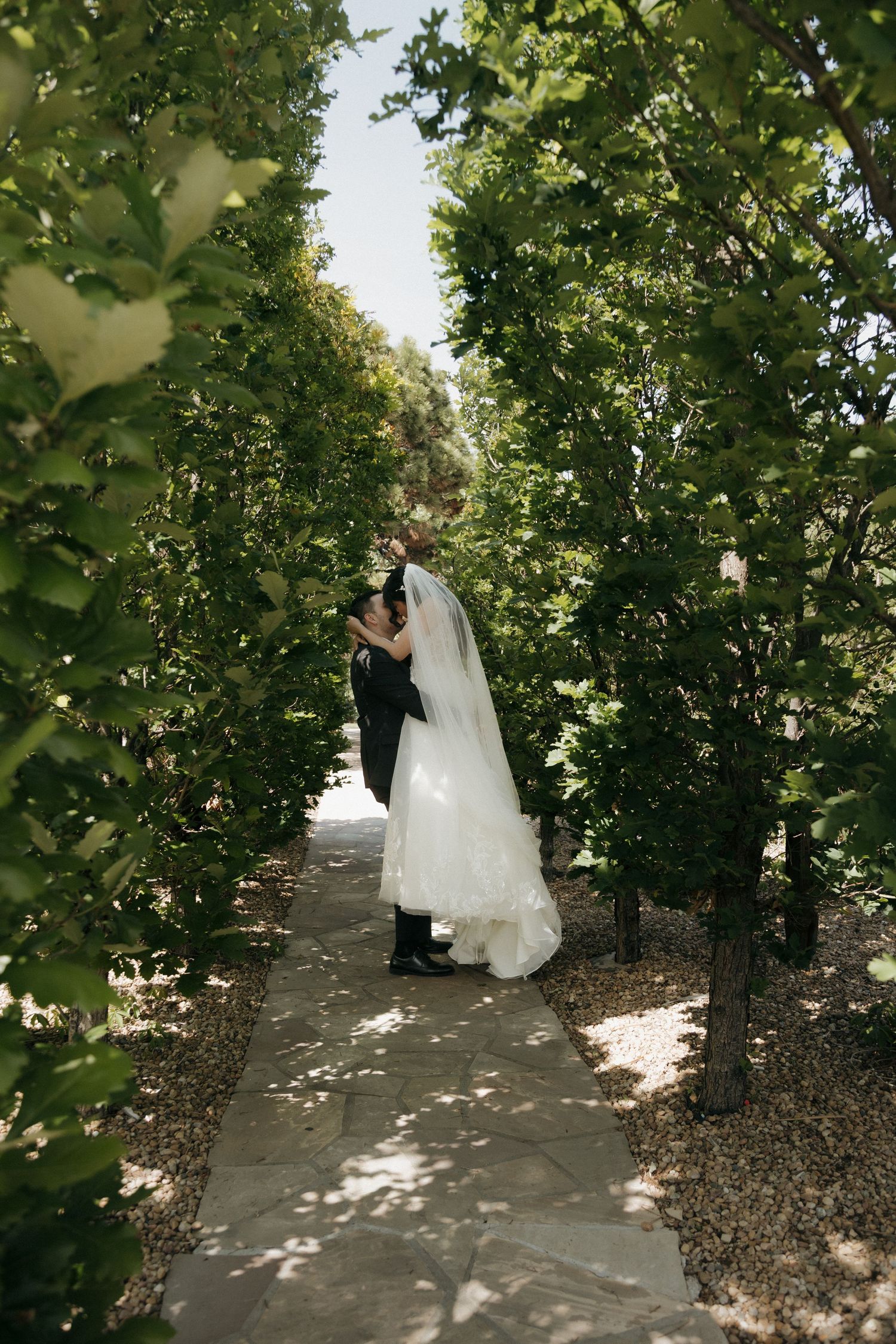 Bride and groom kissing on a stone pathway surrounded by tall green trees.